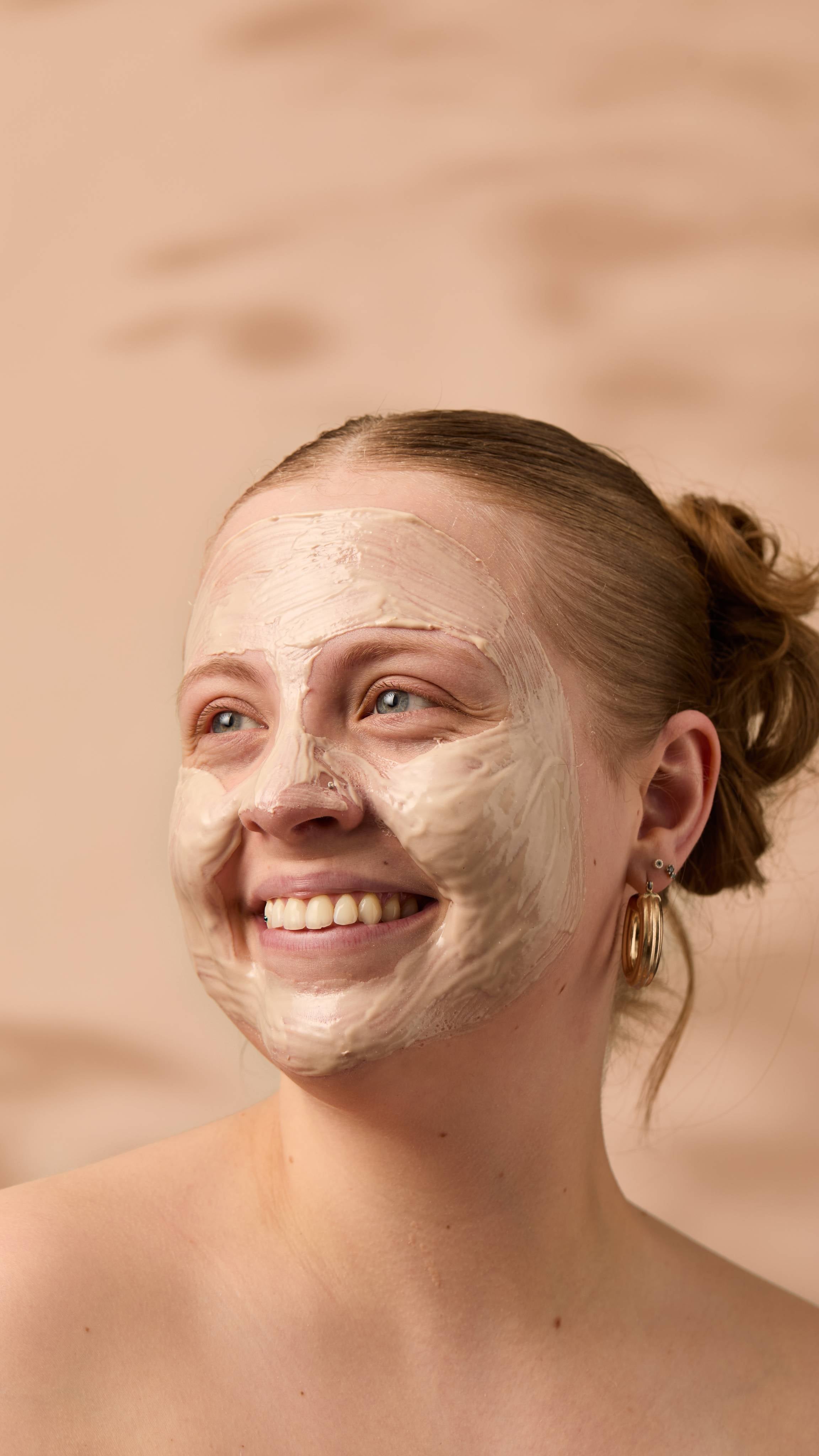 An image of the model's face on a warm, beige background. Their copper hair is tied back, and they have applied the thick Coconut Shie face mask over their full face as they smile.