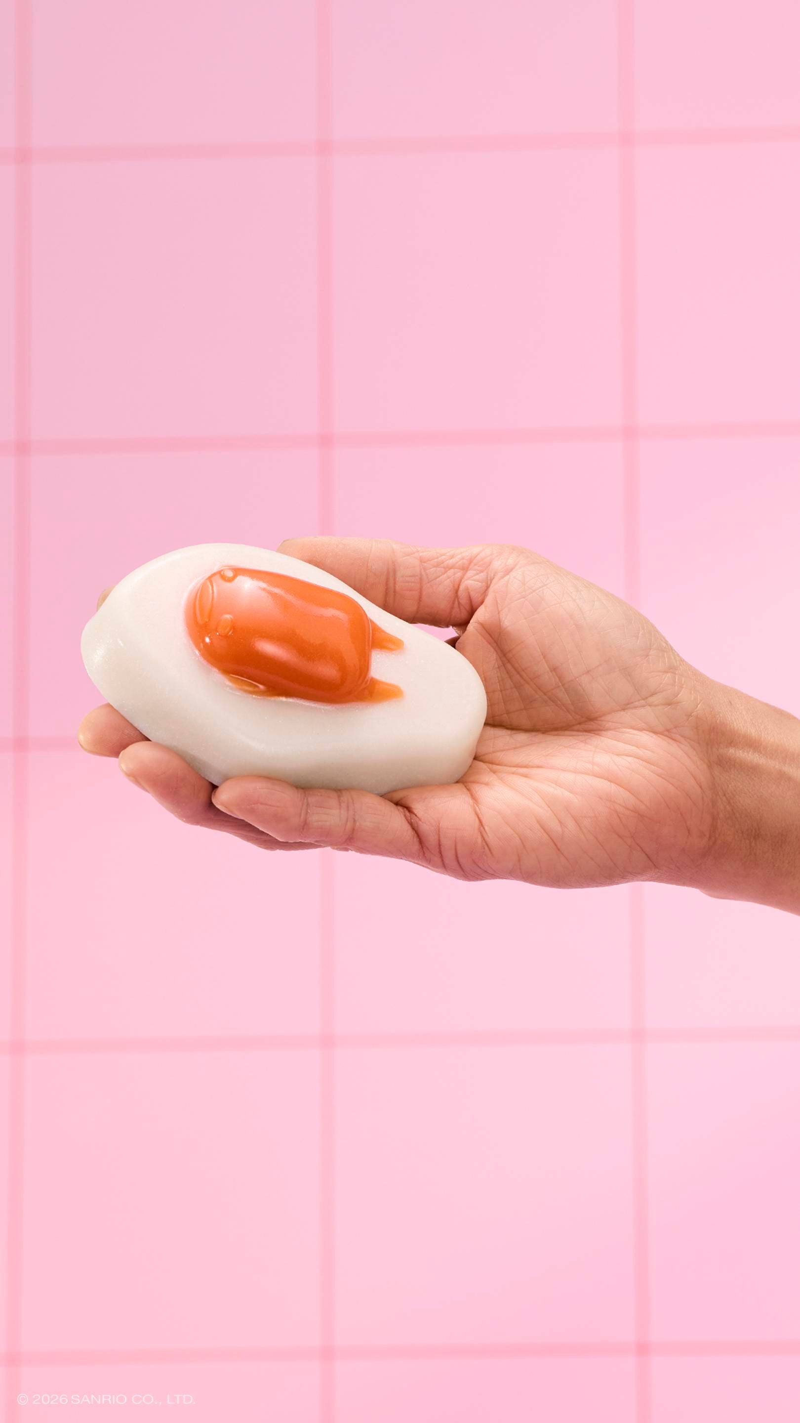 A hand holds a glossy, fried-egg-shaped shower jelly against a background of pink square tiles. The character Gudetama is featured as the orange "yolk" on the white jelly base.