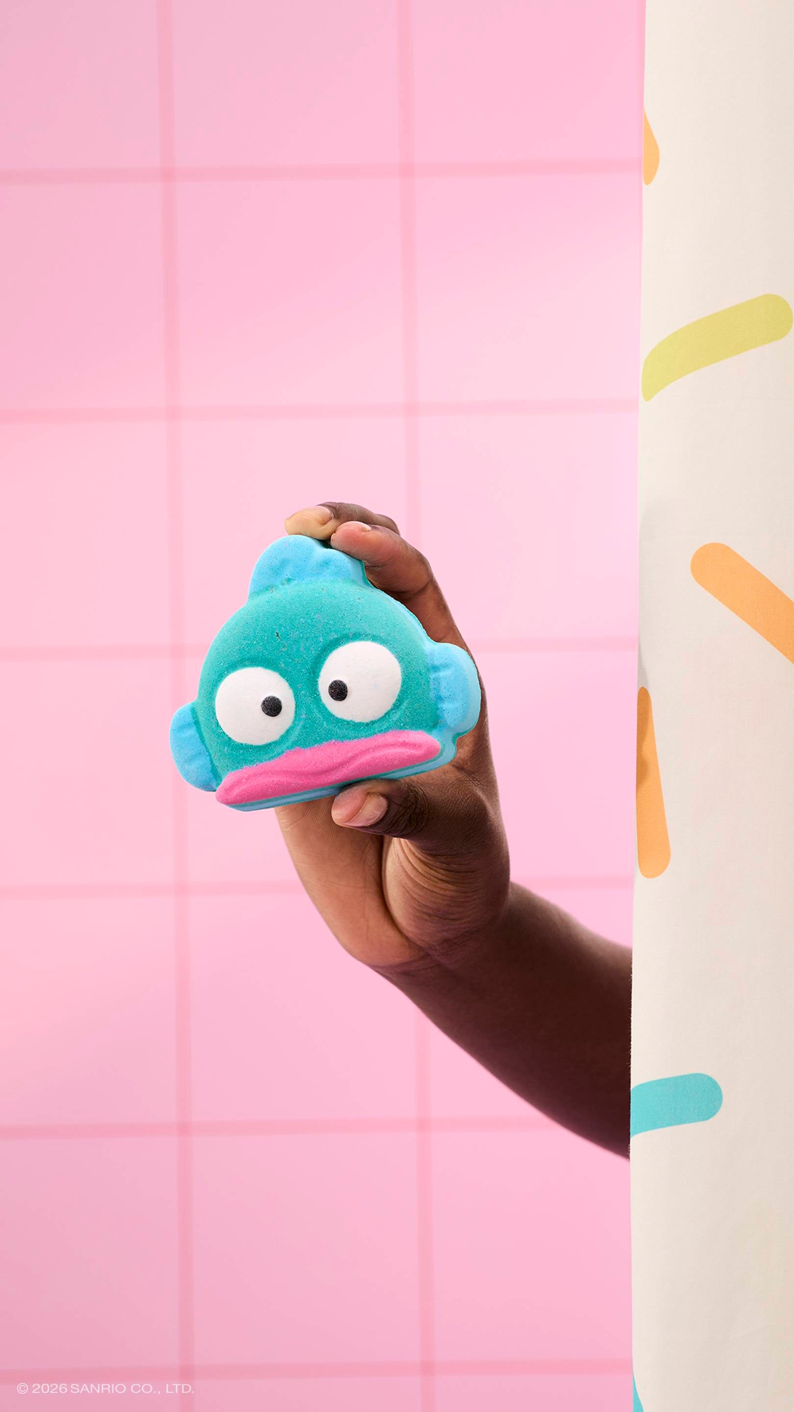 A hand holds a teal, fish-shaped bath bomb with a pink mouth and large white eyes. The person is partially hidden behind a sprinkle-patterned shower curtain, set against a background of pink square tiles.