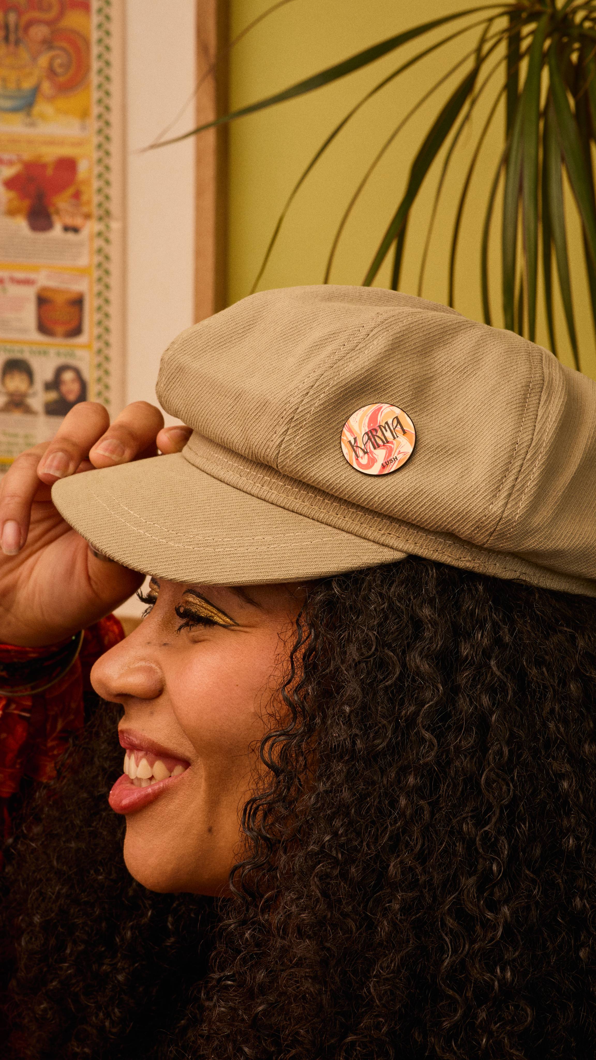 The image shows a person with curly dark hair smiling, wearing a beige newsboy cap. The Karma pin badge, with its red, orange, and white marbled design, is pinned on the cap. A blurred indoor scene with greenery is in the background.