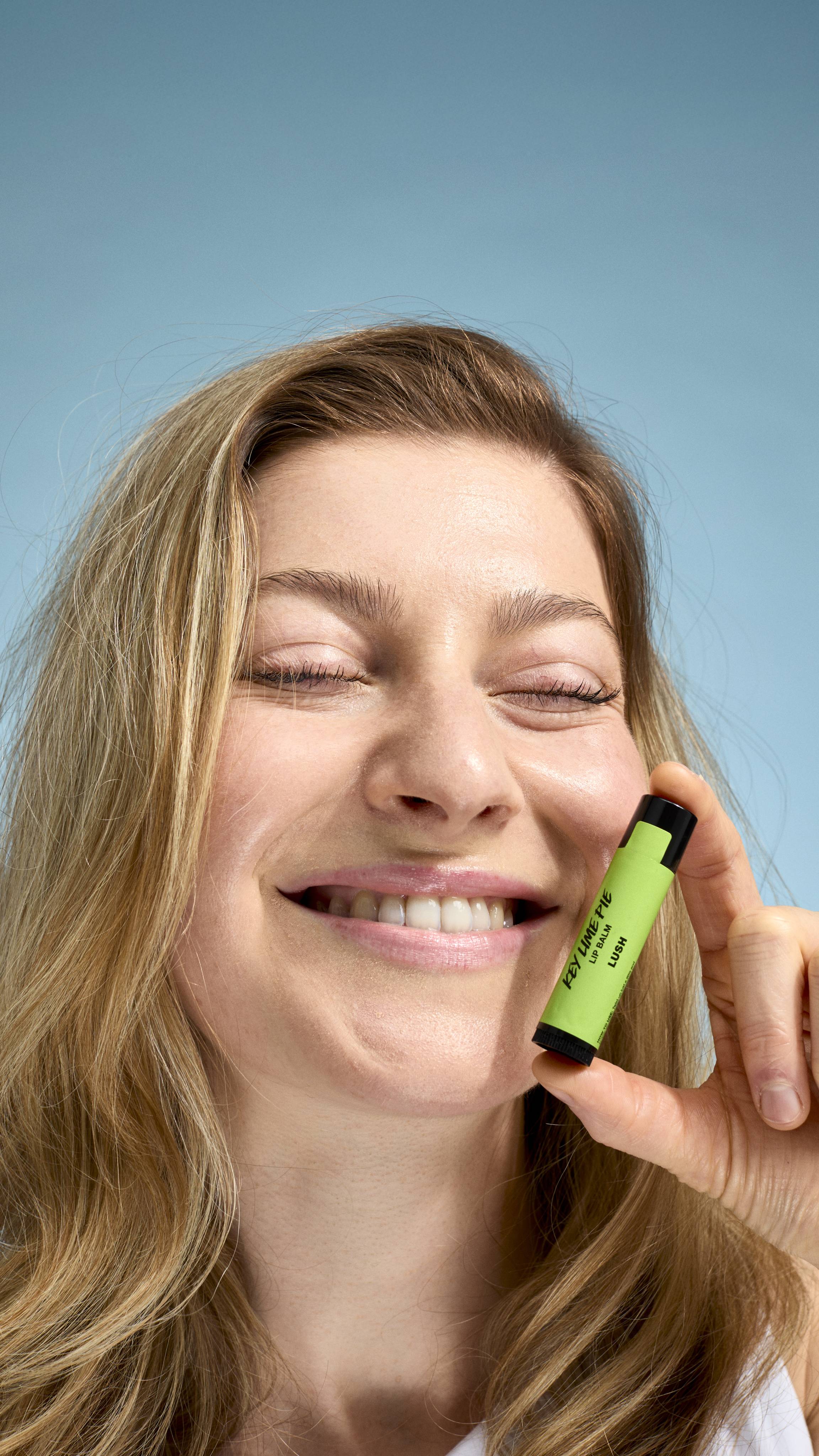 A close-up of a model with fair skin and dark blonde hair, smiling with their eyes closed. They are holding a bright green-labelled tube of Lush 'Key Lime Pie' lip balm against their cheek. The background is a soft, light blue.