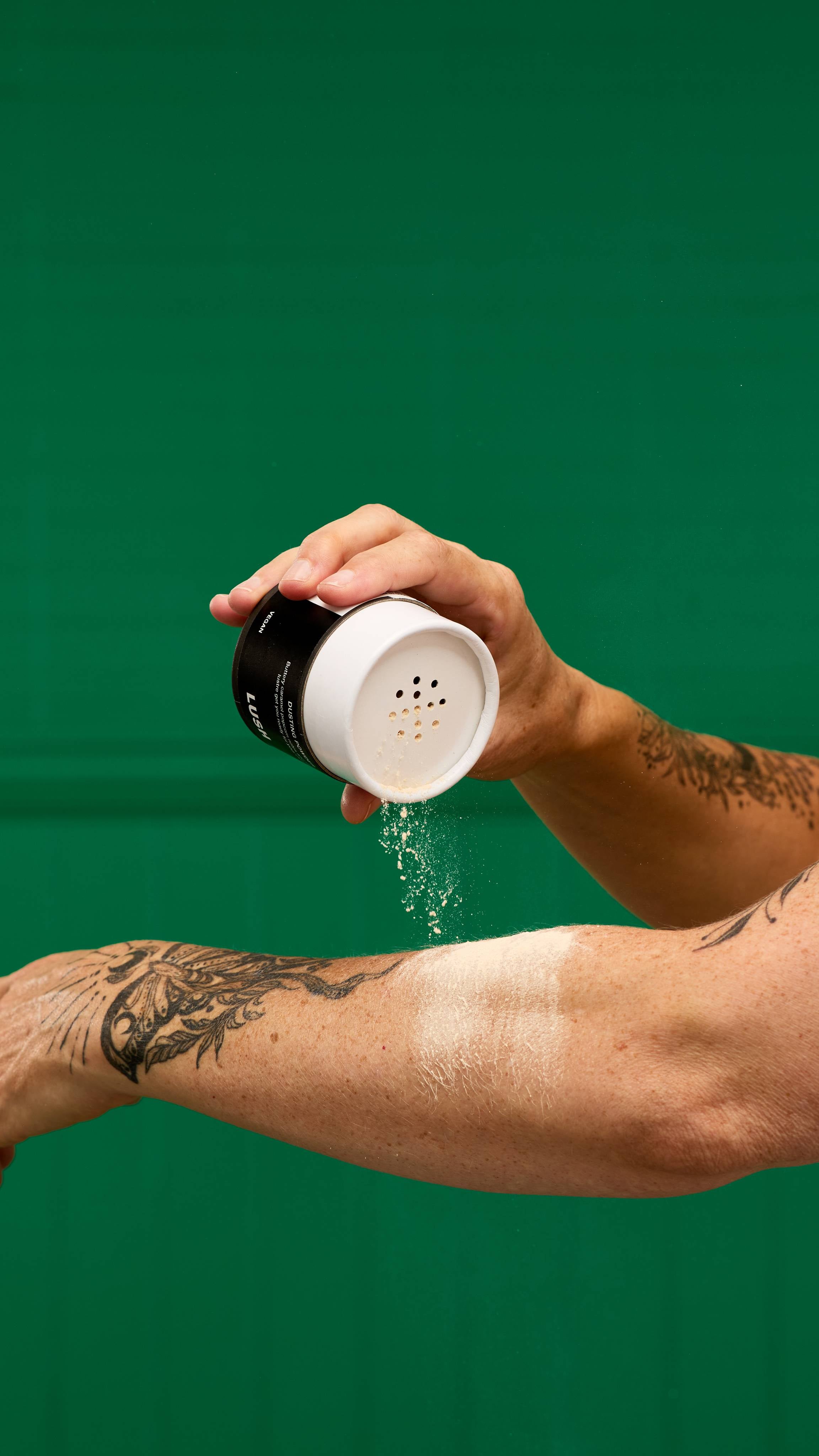A person's hand shakes a LUSH Let the Good Times Roll dusting powder container, with a fine cloud of white powder falling onto their tattooed forearm. The background is a vibrant green.