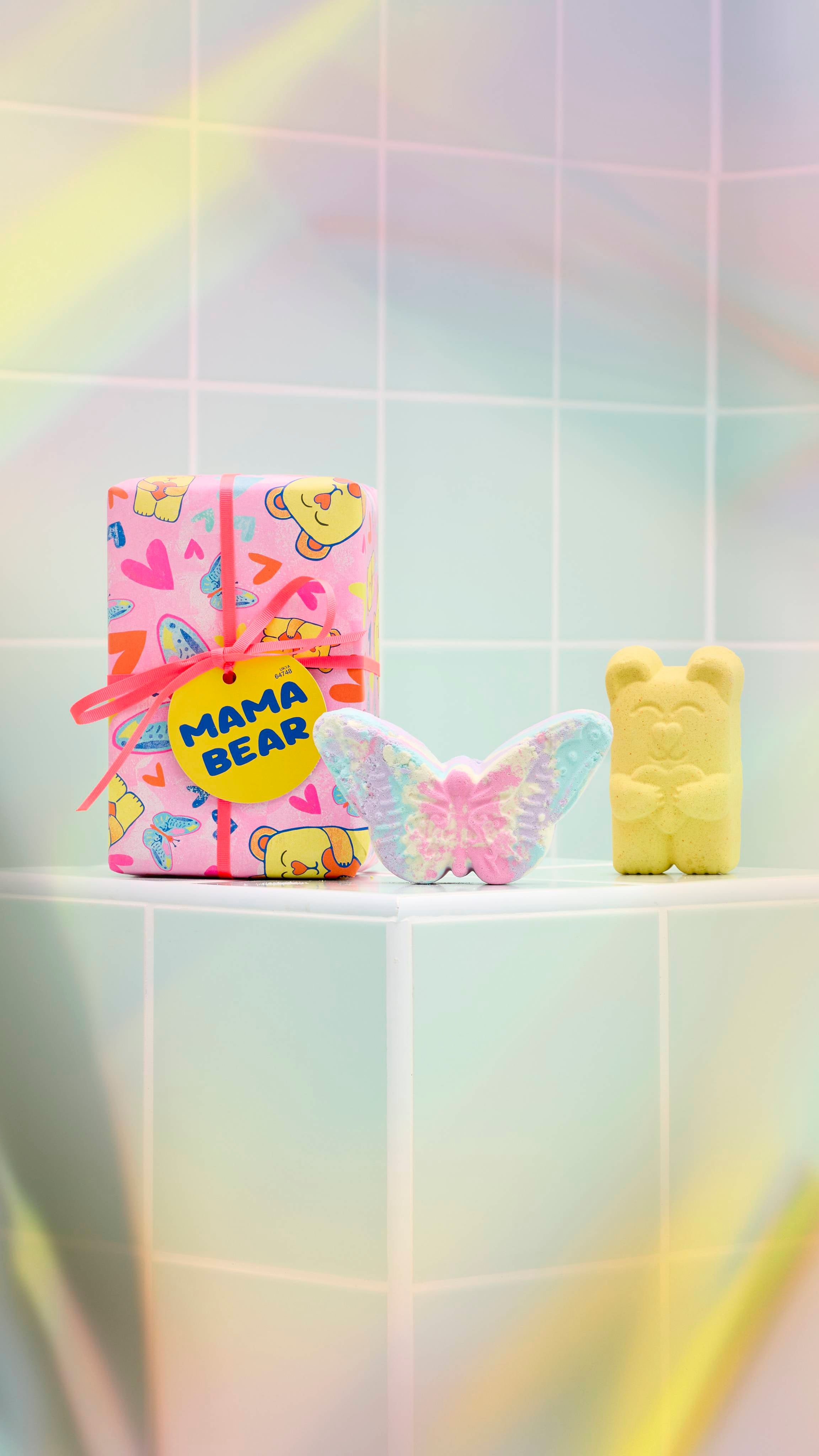 The image shows a brightly wrapped Lush "Mama Bear" gift standing on a pale green tiled ledge. Beside it are a pastel-coloured butterfly bath bomb and a yellow bear-shaped bath bomb.