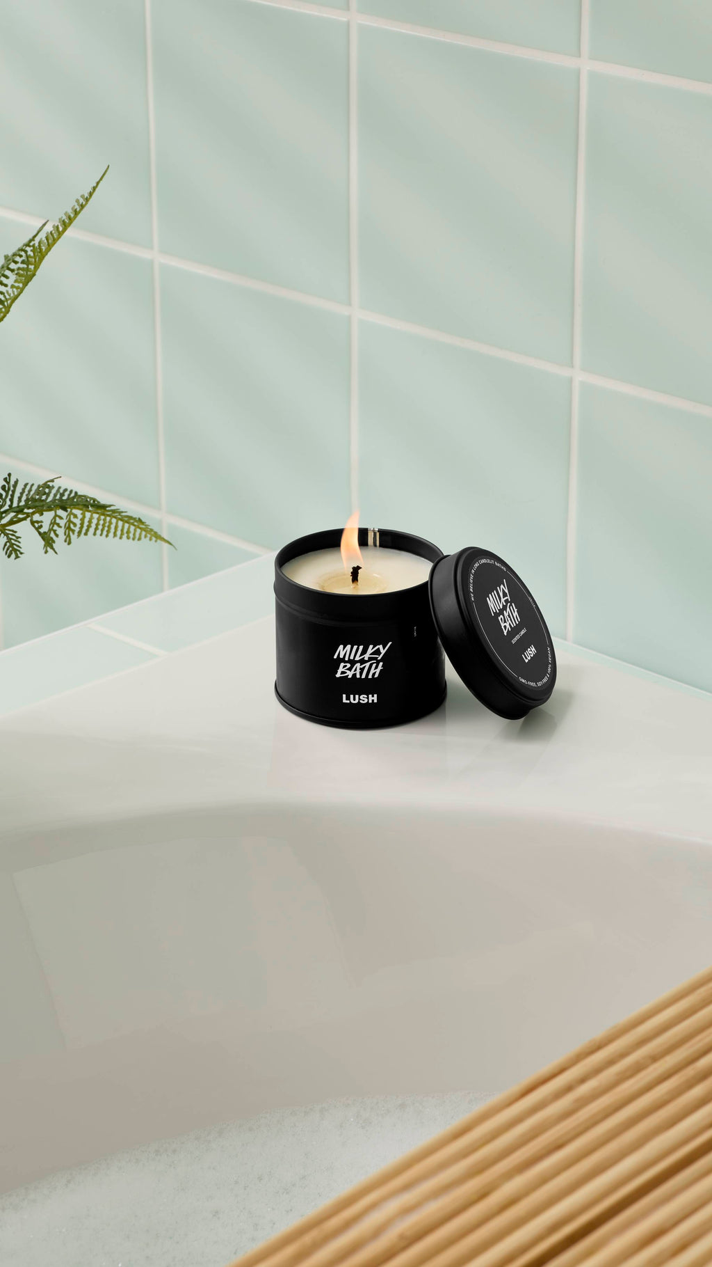 An open, lit Milky Bath candle in a matte black tin rests on the corner of a white bathtub. The lid is propped up against the container. The background features light mint green wall tiles and the leaves of a green fern.