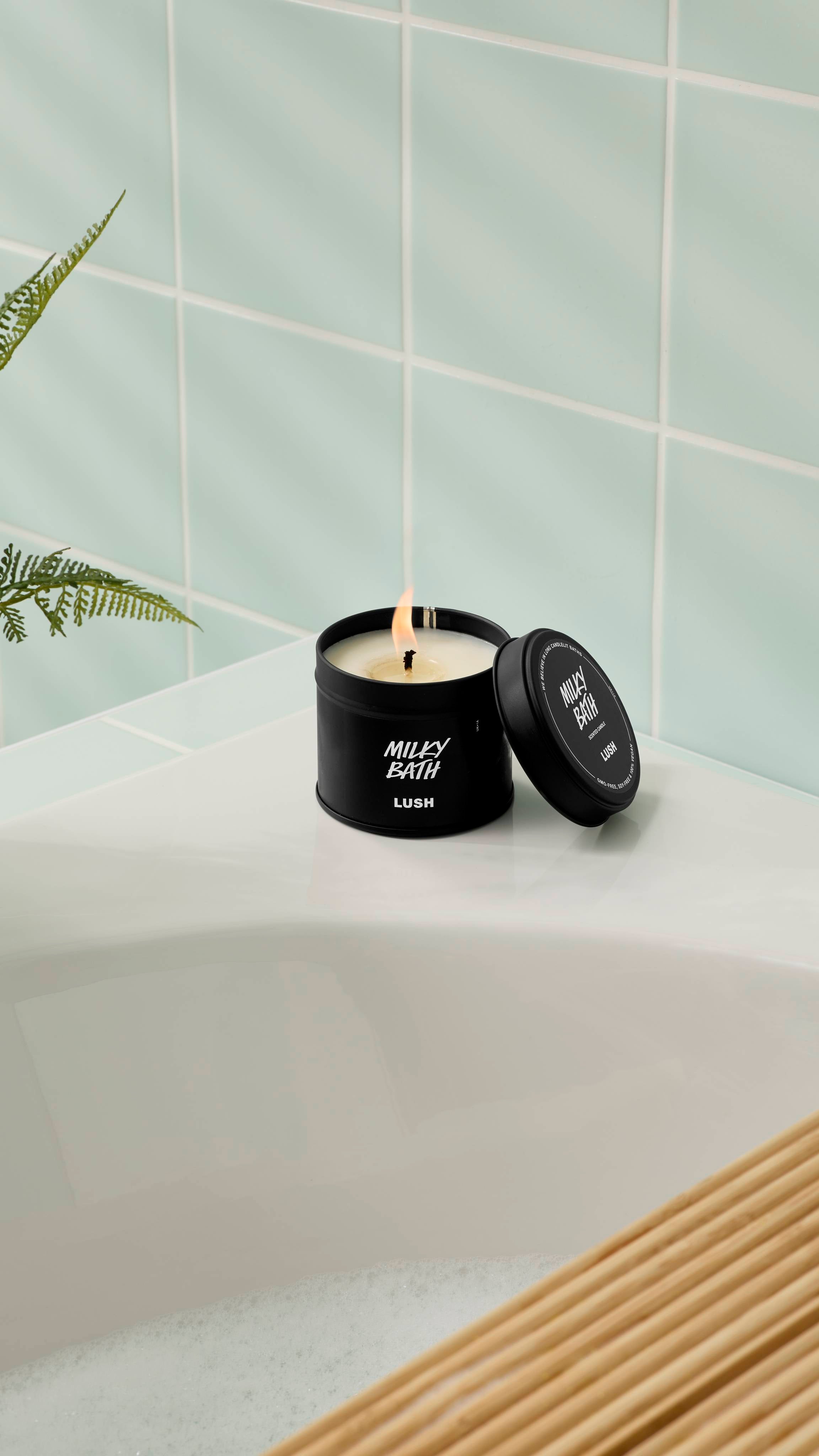 An open, lit Milky Bath candle in a matte black tin rests on the corner of a white bathtub. The lid is propped up against the container. The background features light mint green wall tiles and the leaves of a green fern.