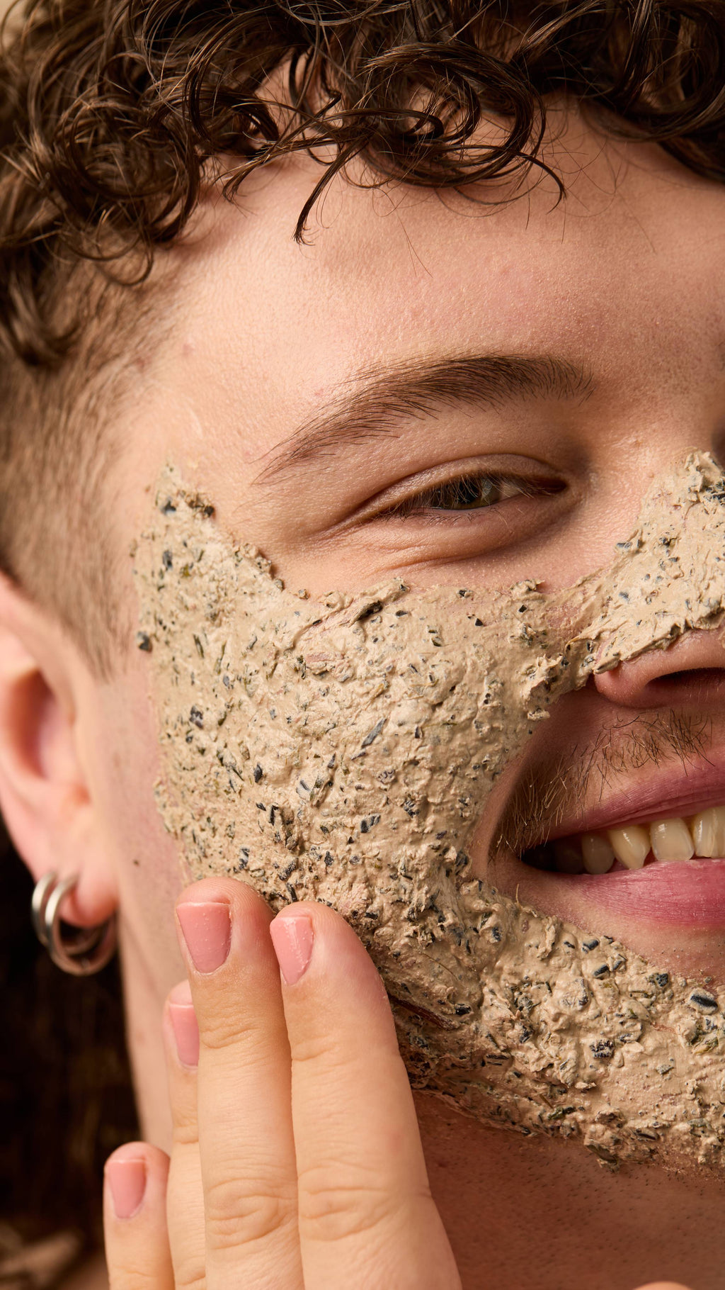 A close-up of the model's face as they have applied the thick textured, Mineralise Me face mask over their full face as they smile.