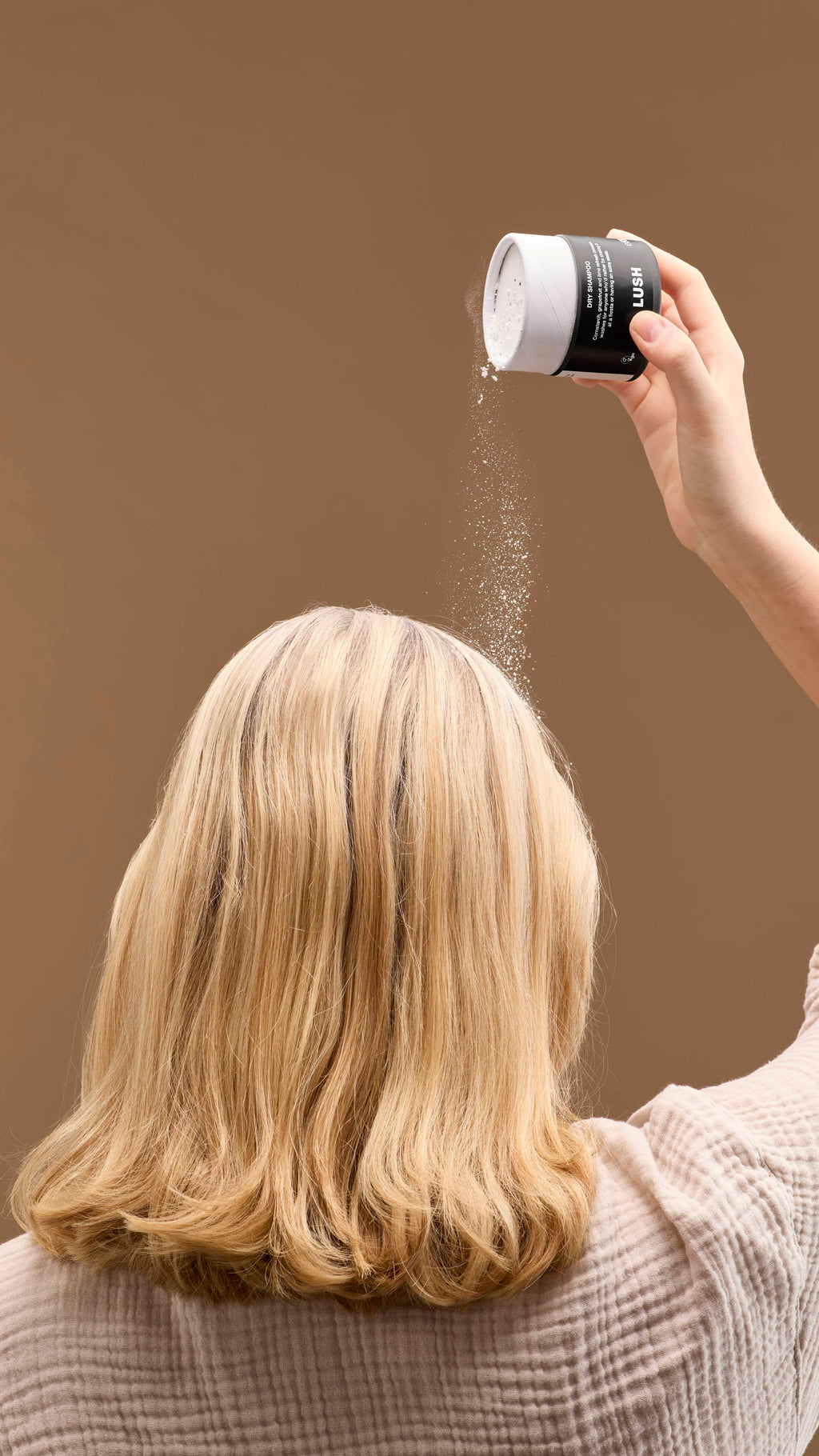 The image shows a hand shaking a cardboard tub of 'No Drought' dry shampoo powder over a person's head. Fine white powder falls onto their light blonde hair, against a solid beige background.