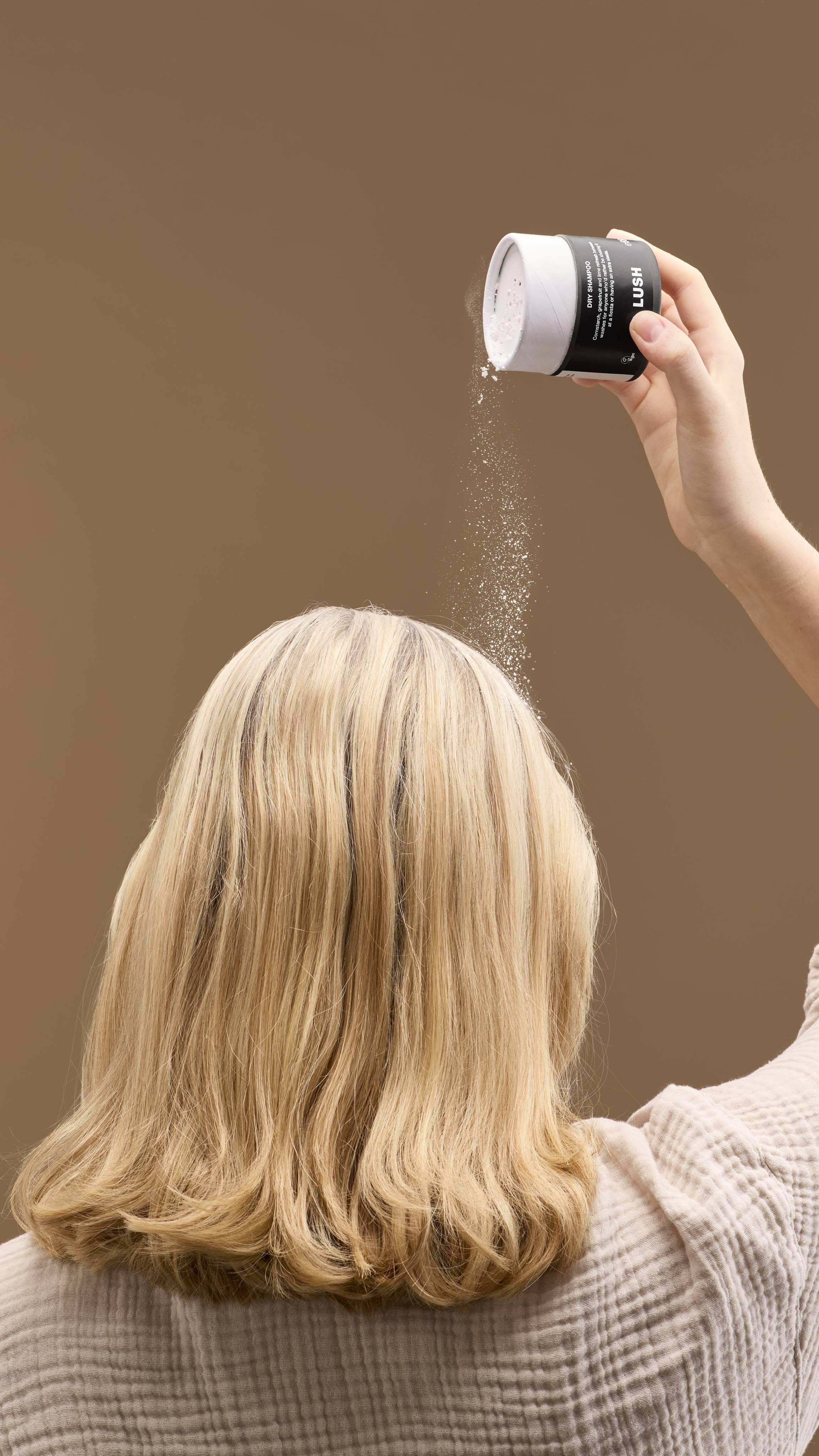 The image shows a hand shaking a cardboard tub of 'No Drought' dry shampoo powder over a person's head. Fine white powder falls onto their light blonde hair, against a solid beige background.