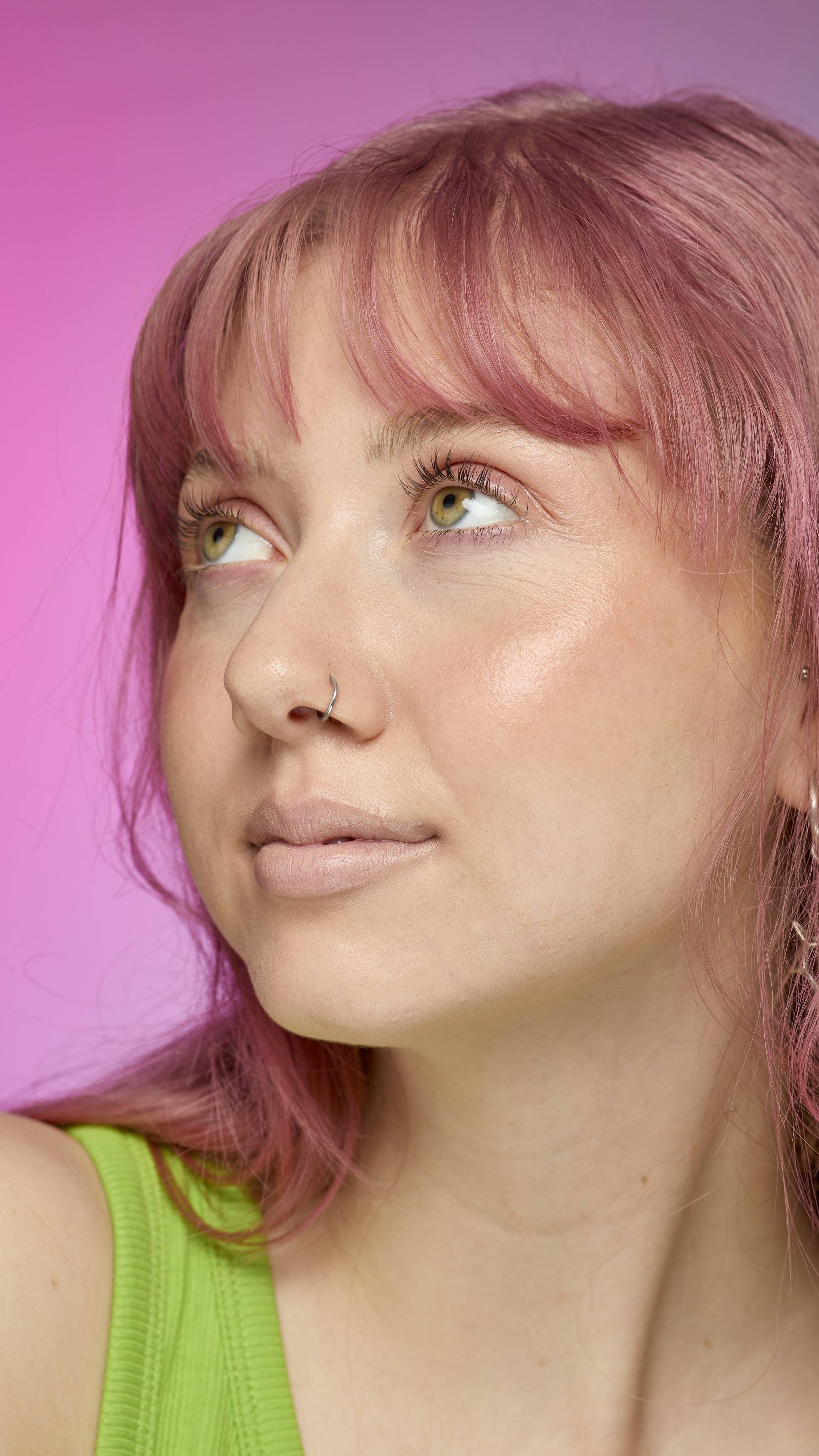 The image shows a fair-skinned model on a pink and blue gradient background. They have pink hair and subtle, peach-nude shimmering lips from using the Peach parfait lip cream.
