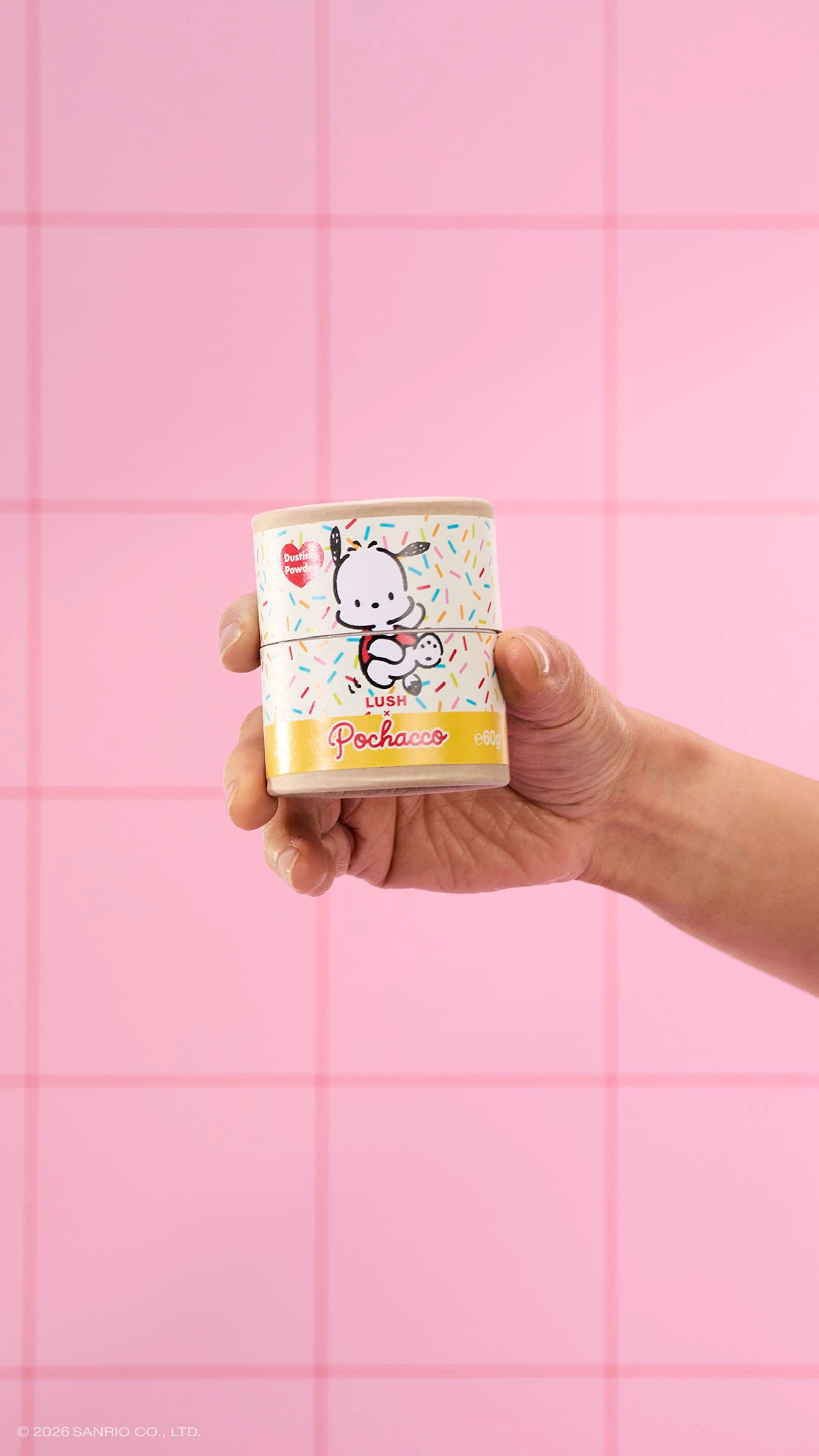 A close-up of a model's hand holding a cardboard tube of Lush x Pochacco dusting powder, featuring a sprinkle pattern and a cartoon dog. The background consists of pink square tiles.