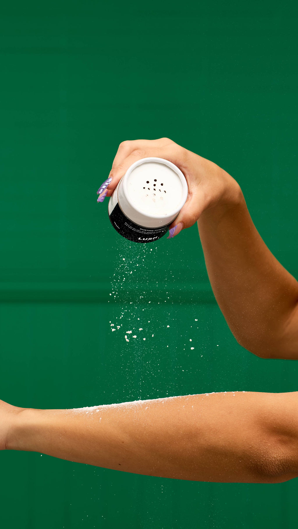 A person's hand with purple-tipped nails shakes a LUSH Sticky Dates dusting powder container, with a fine cloud of white powder falling onto their outstretched forearm. The background is a vibrant green.