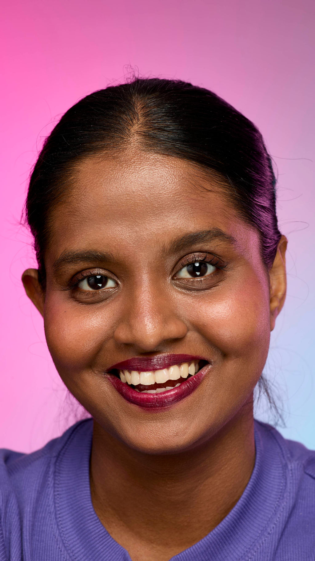 A smiling person with dark hair and skin, wearing a purple top, is shown from the chest up. They are wearing the Lush, deep plum Tartlet lip cream on a pink and purple gradient background.