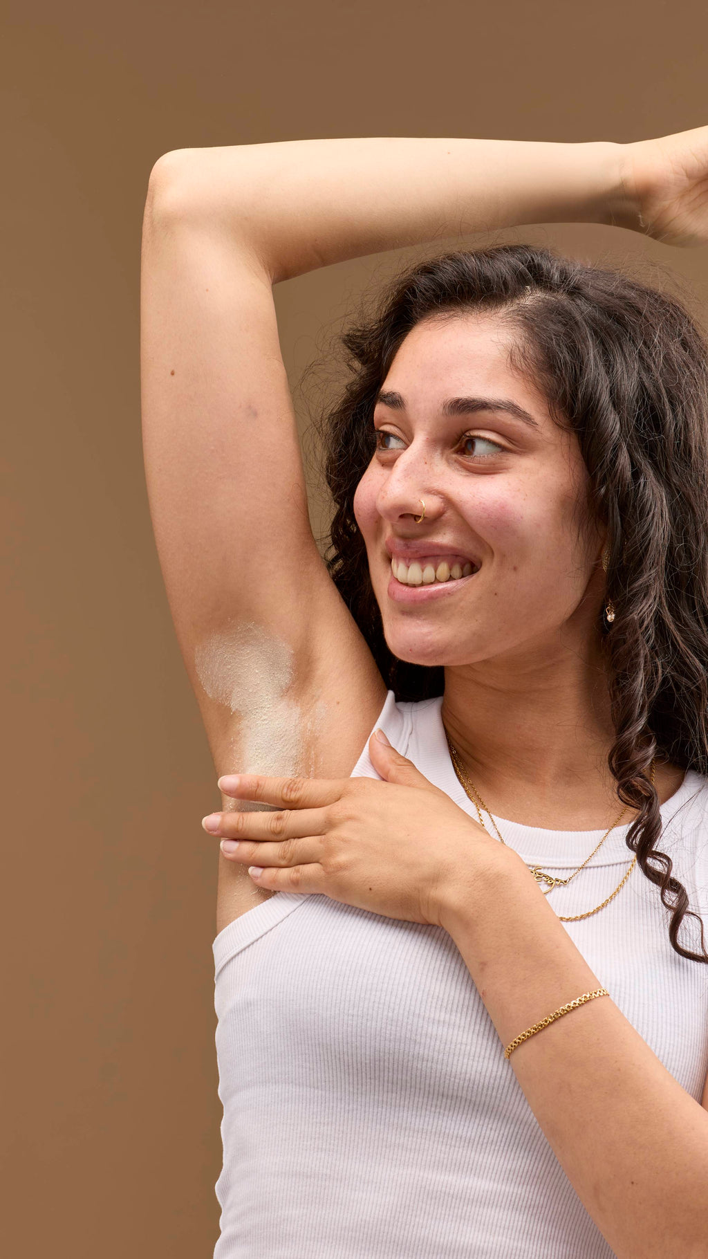 The image shows a person with dark, curly hair, smiling, with a raised arm. A pale cream 'The Greeench' deodorant powder is dusted onto the underarm. The person wears a white vest top and necklaces, against a light beige background.