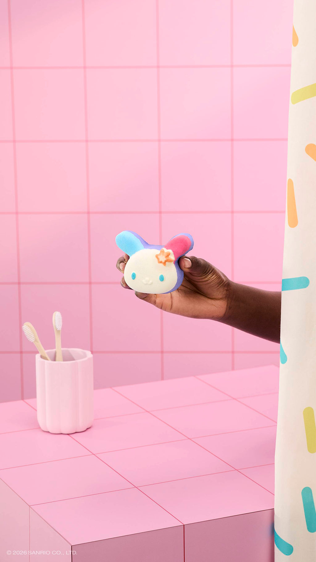 A hand holds a colourful bunny-shaped "U.SA.HA.NA" bath bomb in a pink tiled bathroom. A pink cup with two toothbrushes sits on the counter, and a sprinkle-patterned shower curtain is visible on the right.
