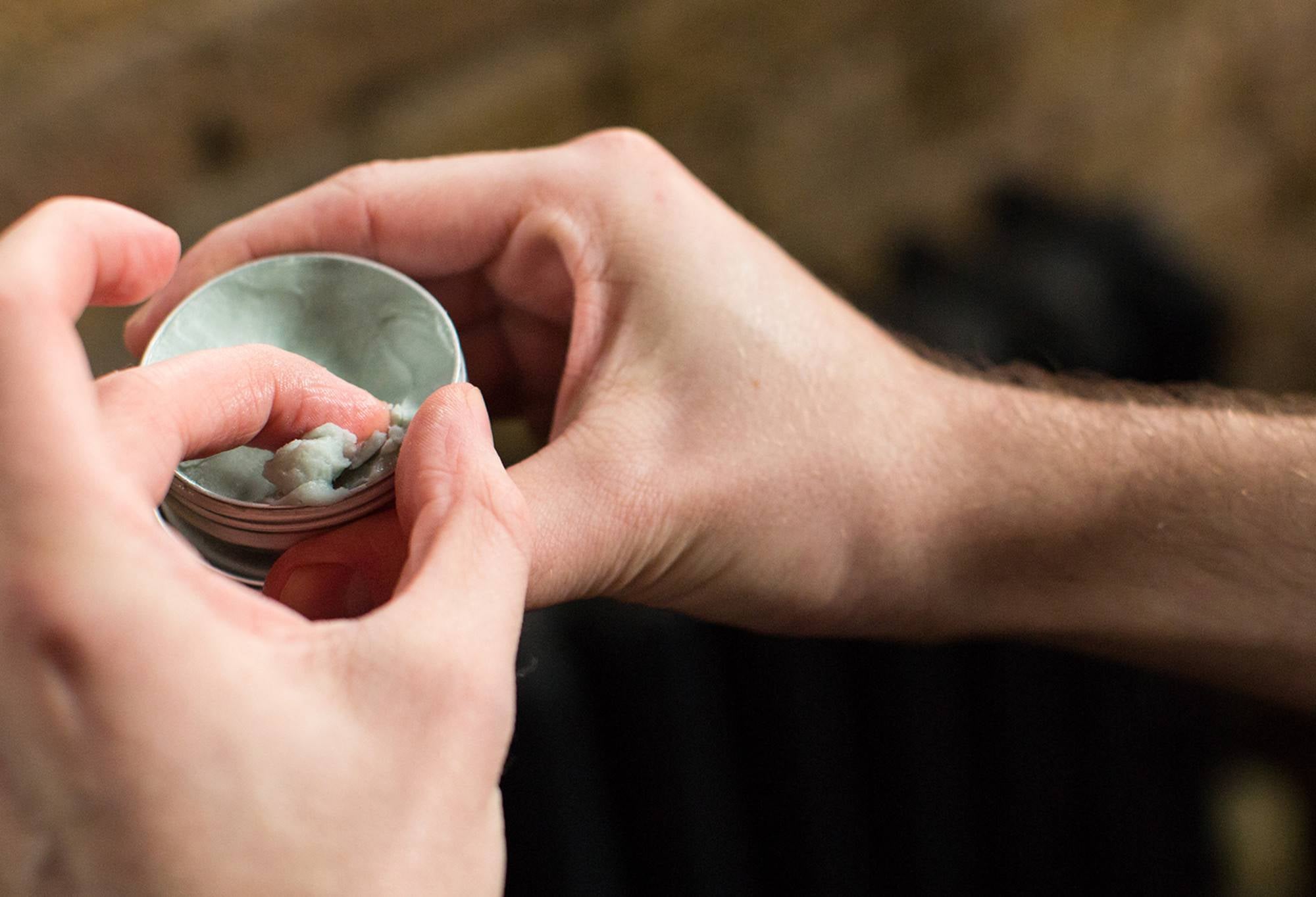 A finger scoops a generous amount of Superbalm, a very thick, balmy, pale green coloured scalp treatment, out of its metal tin.