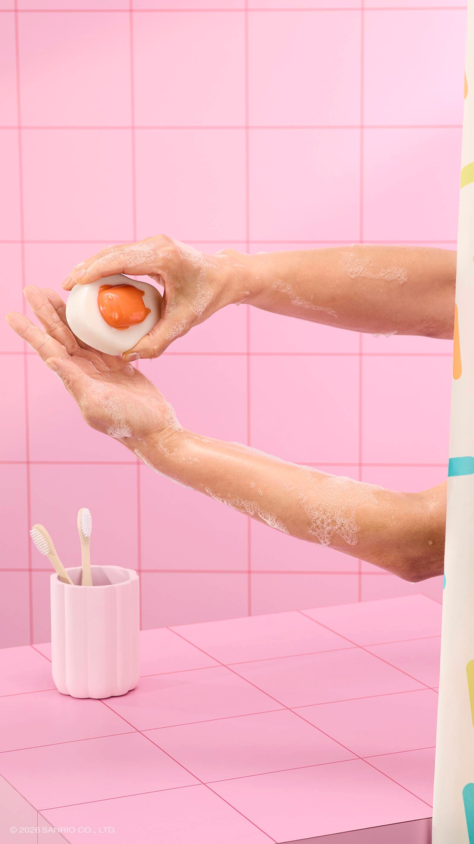 A person lathers their soapy arms using a glossy, egg-shaped "Gudetama" shower jelly in a pink tiled bathroom. A pink toothbrush holder and a sprinkle-patterned curtain are visible.
