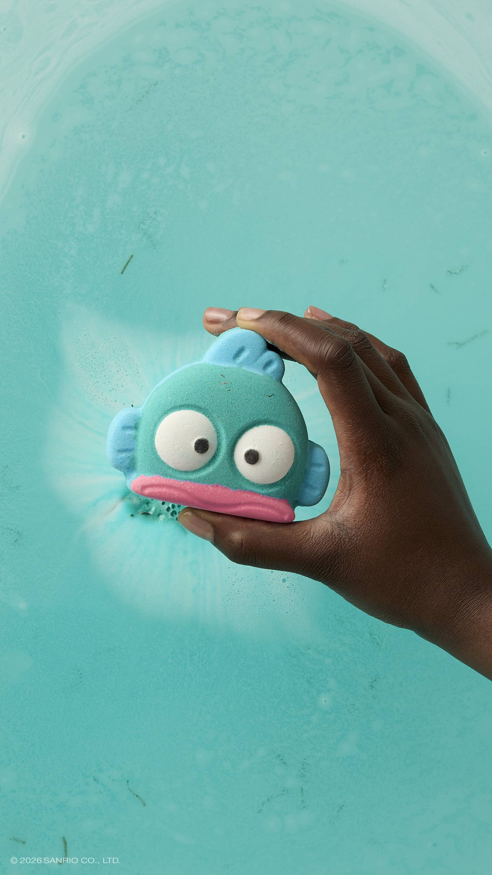 A hand holds a teal, fish-shaped bath bomb over a bath of bright turquoise water. The bath bomb features a pink mouth and large, startled white eyes. A soft white fizz is visible in the water directly behind the product.