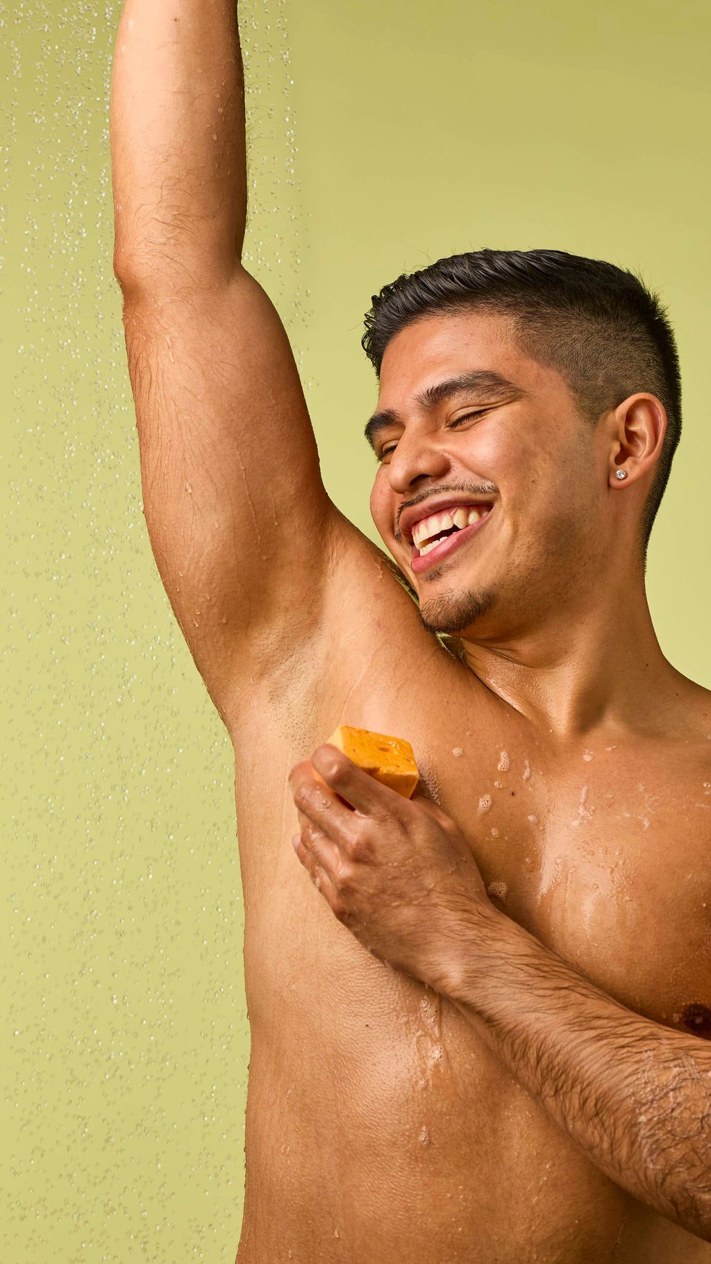 The image shows a person with short dark hair and a wide smile, rubbing the pastel orange Passion Fruit Delight floating soap onto their raised underarm. Water droplets fall on them against a light green background.