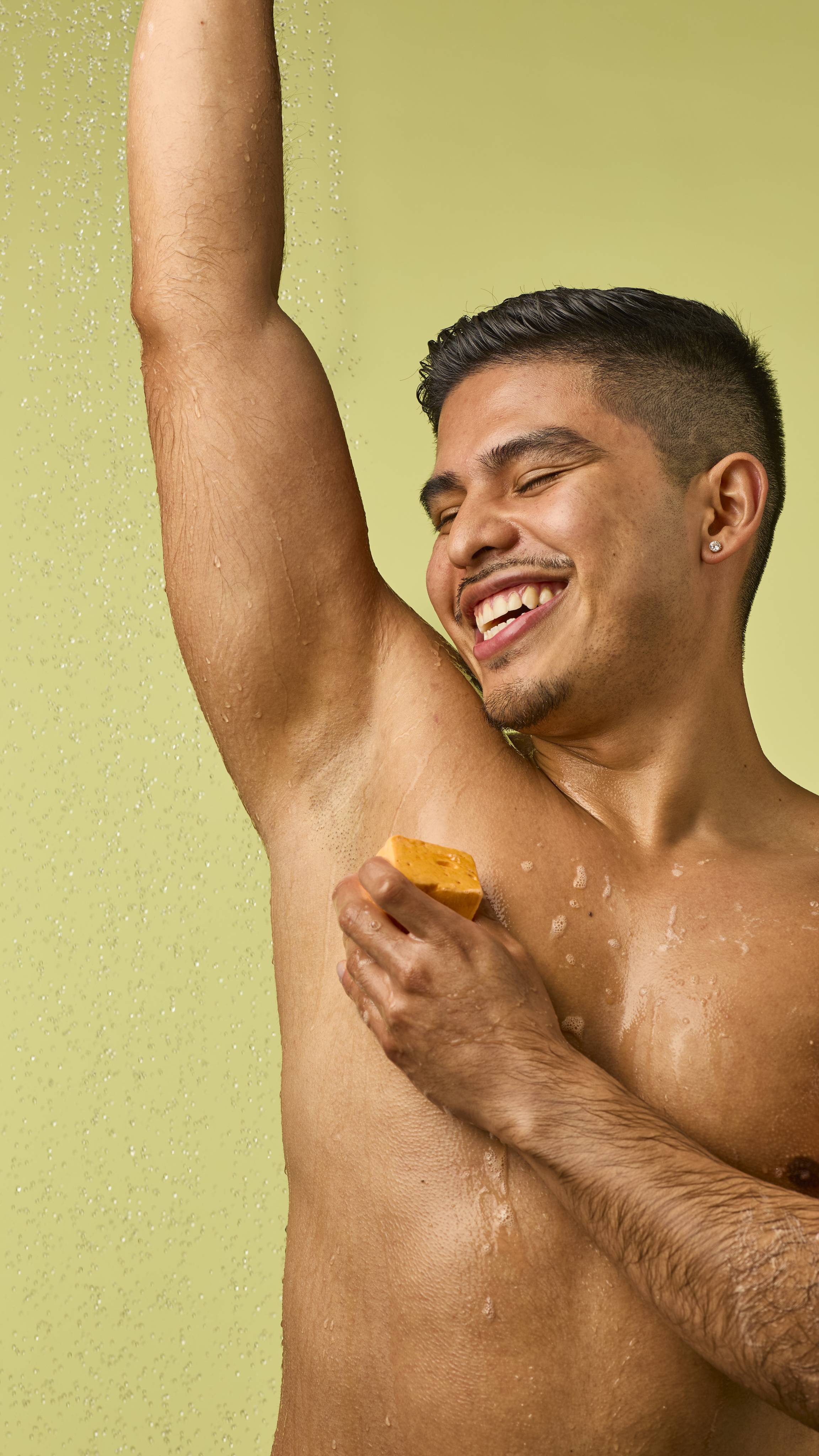 The image shows a person with short dark hair and a wide smile, rubbing the pastel orange Passion Fruit Delight floating soap onto their raised underarm. Water droplets fall on them against a light green background.