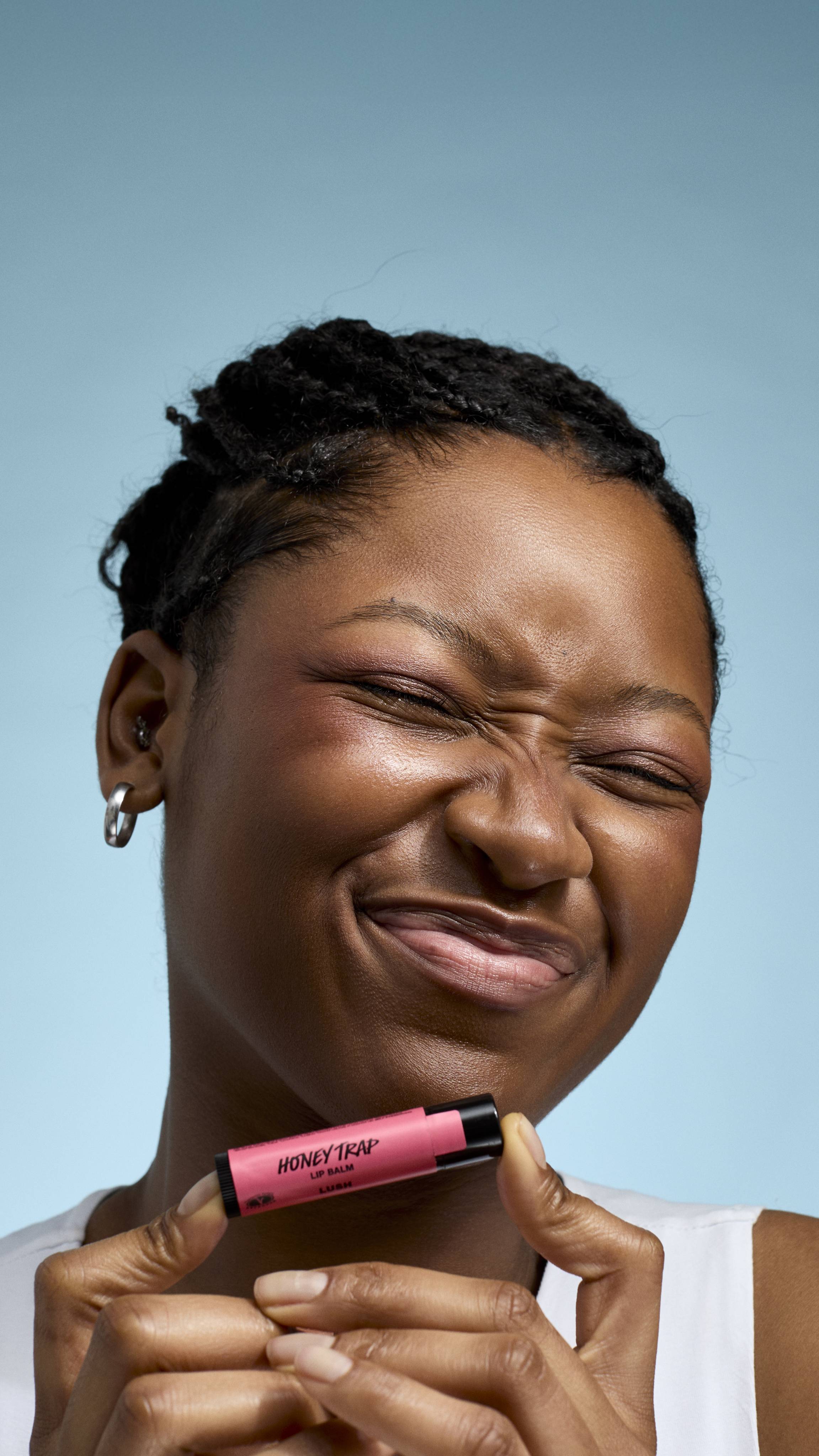 A close-up of a model with deep-toned skin and braided hair, holding a pink tube of Lush 'Honey Trap' lip balm between both hands. They have their eyes closed in a playful, scrunch-faced expression against a soft blue background.