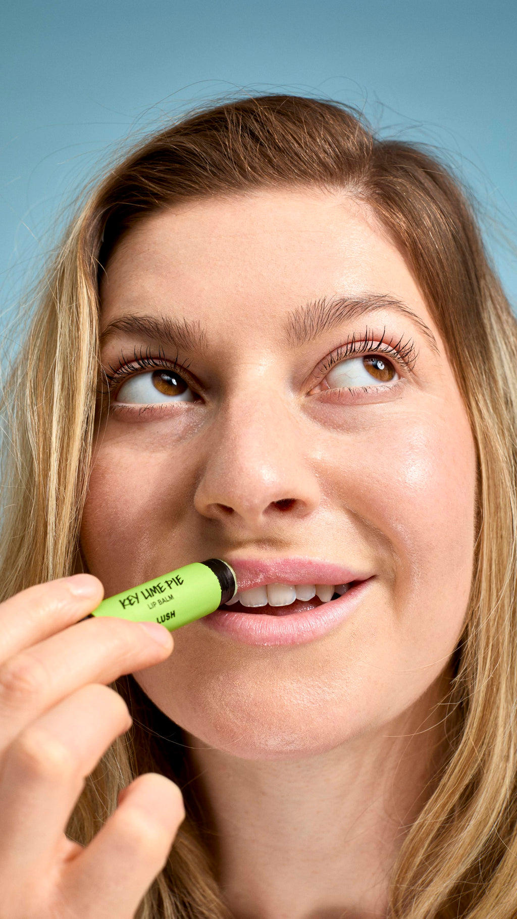 A close-up of a model applying 'Key Lime Pie' lip balm to their lips. They have a natural smile and are looking upwards and to the side, holding the bright green-labelled Lush tube against their mouth against a soft, light-blue background.