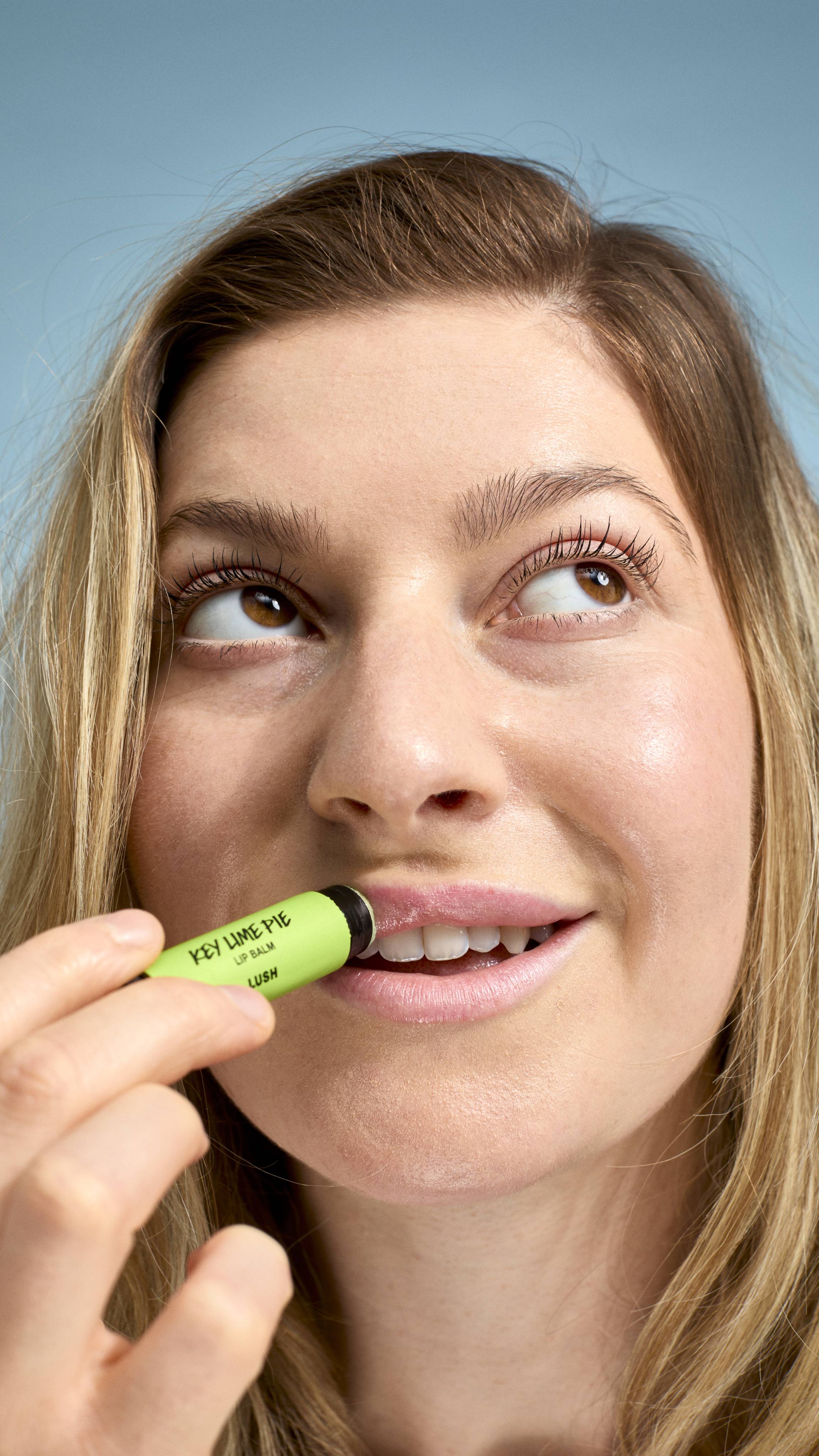 A close-up of a model applying 'Key Lime Pie' lip balm to their lips. They have a natural smile and are looking upwards and to the side, holding the bright green-labelled Lush tube against their mouth against a soft, light-blue background.