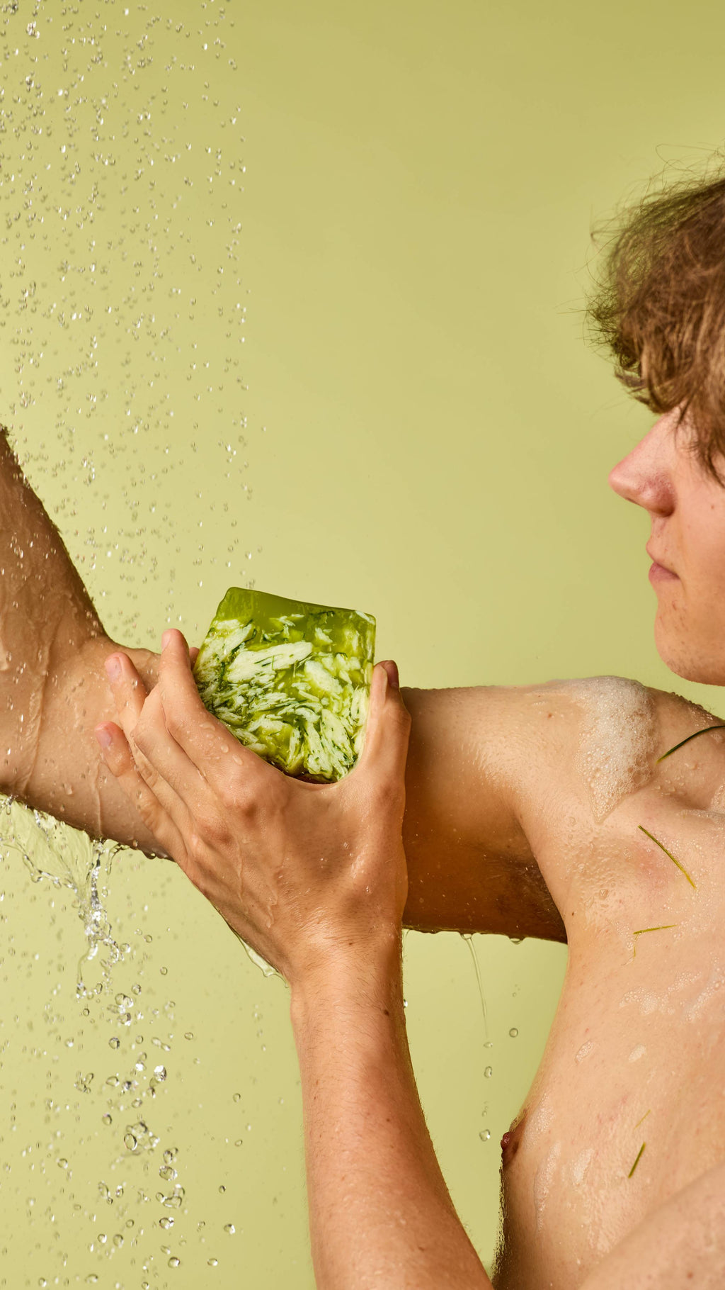 The image shows a person with short brown hair rubbing an off-square slice of translucent green and white Lemongrass soap onto their arm. Water droplets fall on their skin against a light green background.