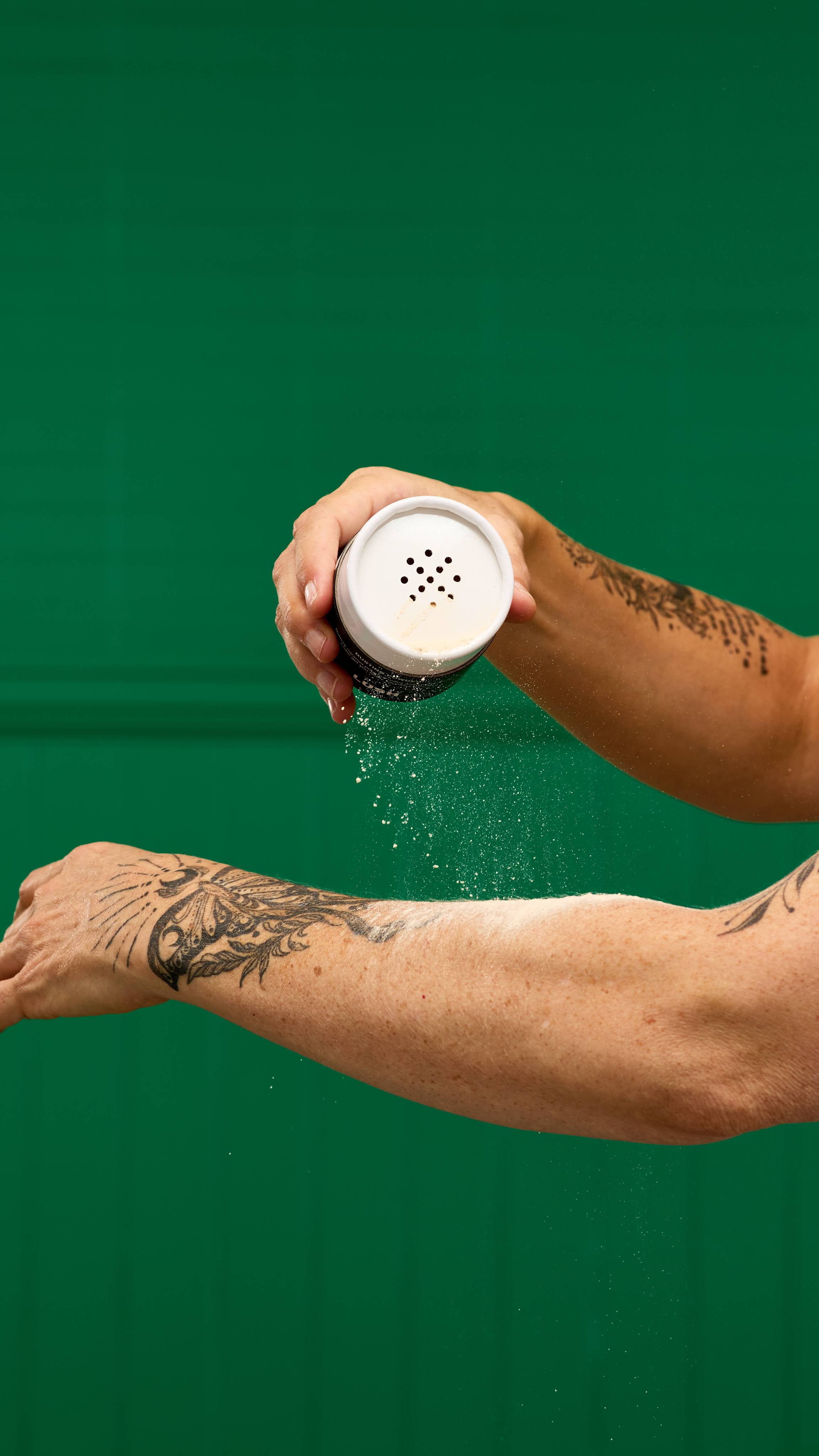 A person's hand shakes a LUSH Let the Good Times Roll dusting powder container, with a fine cloud of white powder falling onto their tattooed forearm. The background is a vibrant green.