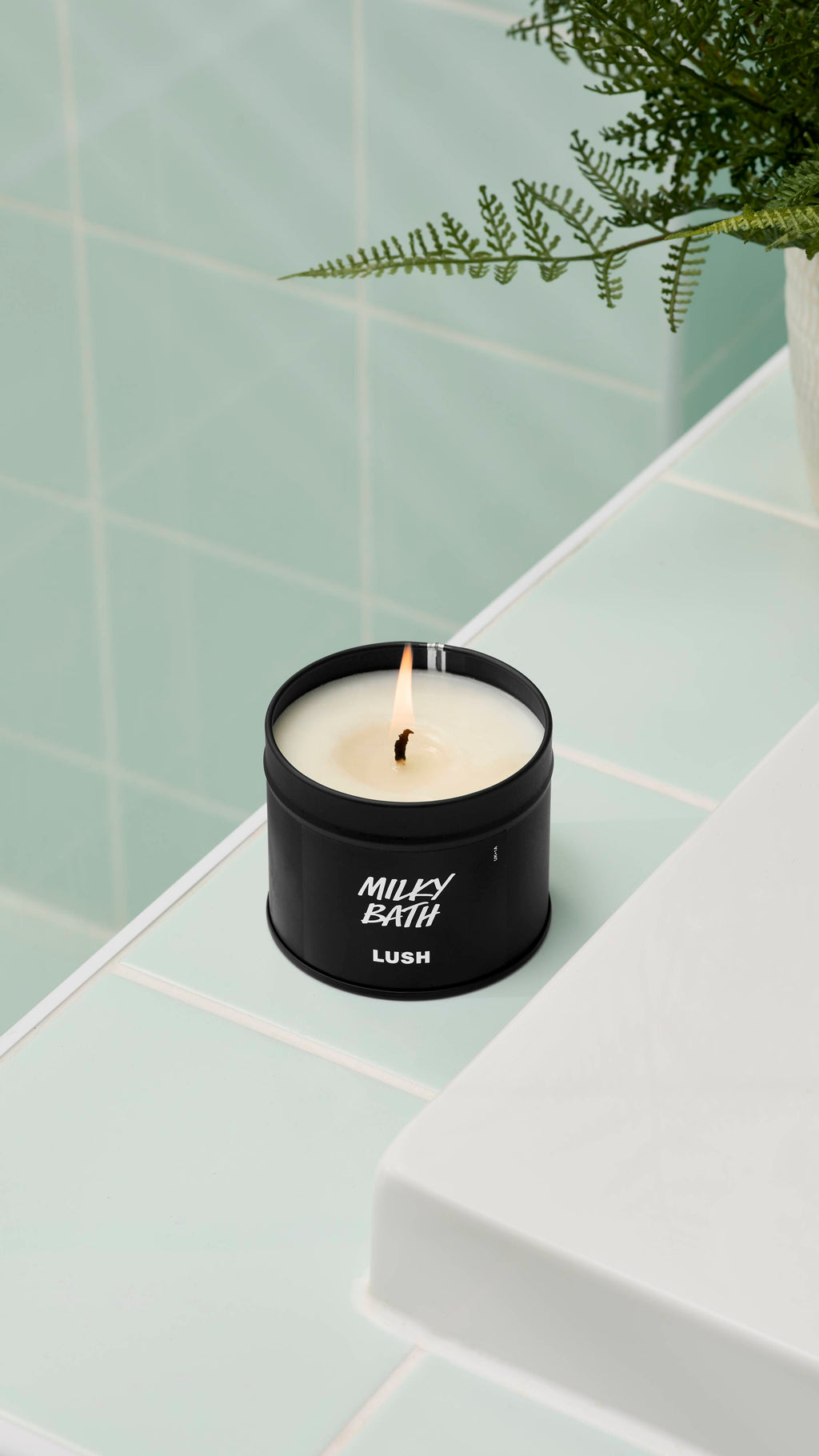A close-up of an open, lit Milky Bath candle by Lush in a matte black tin, with the lid propped against the side. It sits on the edge of a white bathtub. Mint green tiles and a fern are in the background. All glitter used is plastic-free.