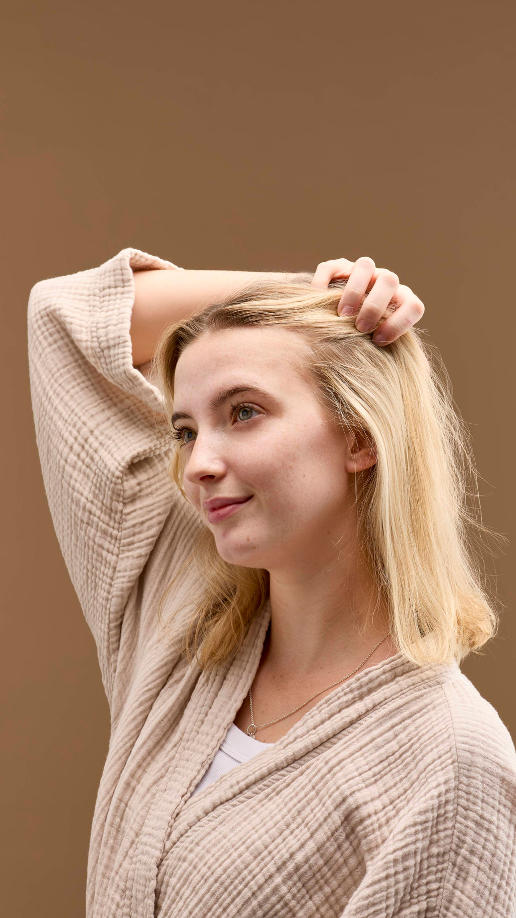 The image shows a person with light blonde hair, smiling. They run a hand through their hair, which appears clean and refreshed from the No Drought dry shampoo. They wear a light-coloured robe, against a solid beige background.