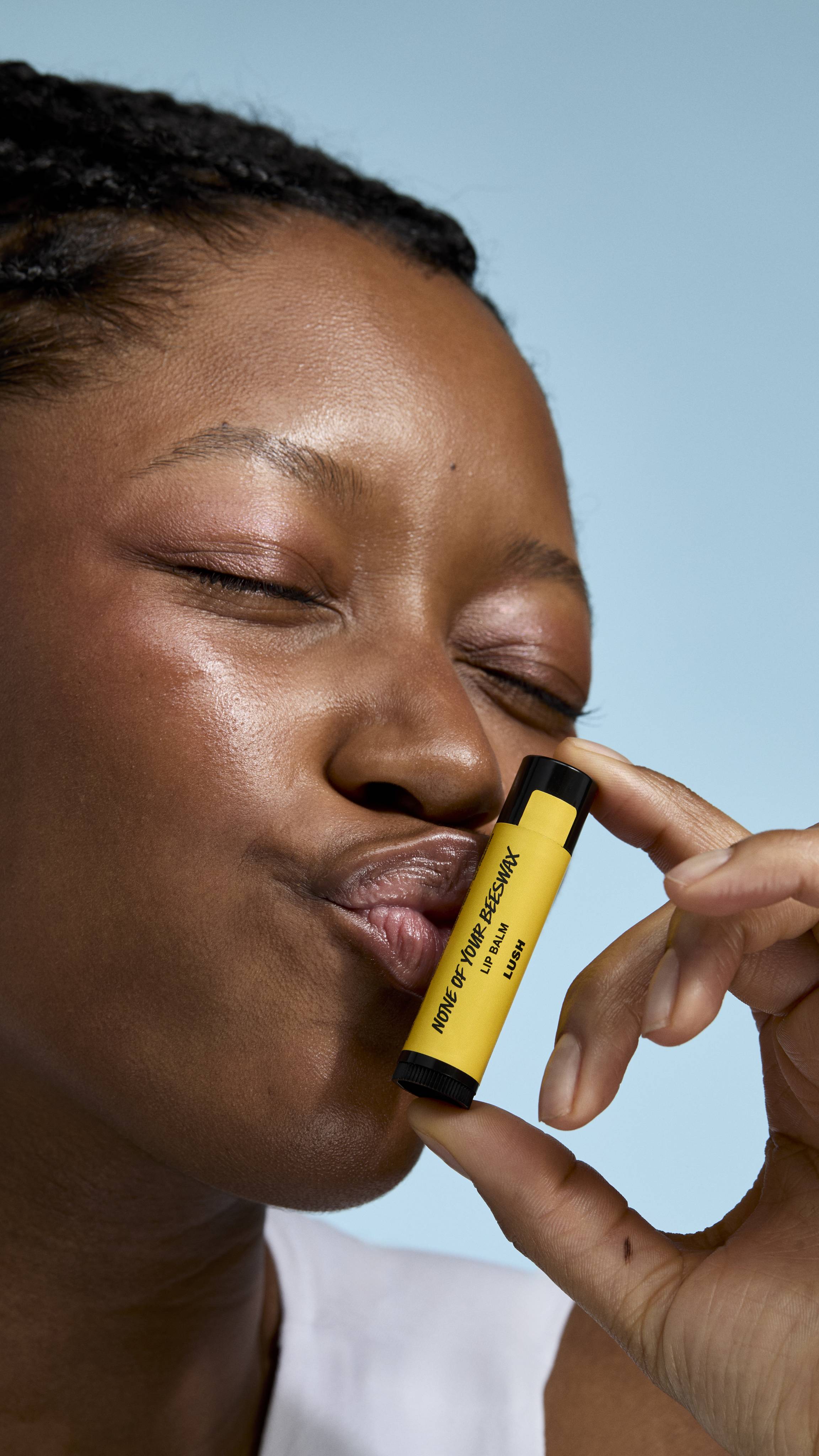 A close-up of a model with deep-toned skin and braided hair holding a yellow 'None of Your Beeswax' lip balm. They have their eyes closed and are making a playful kissing face, with their lips touching the tube against a soft blue background.