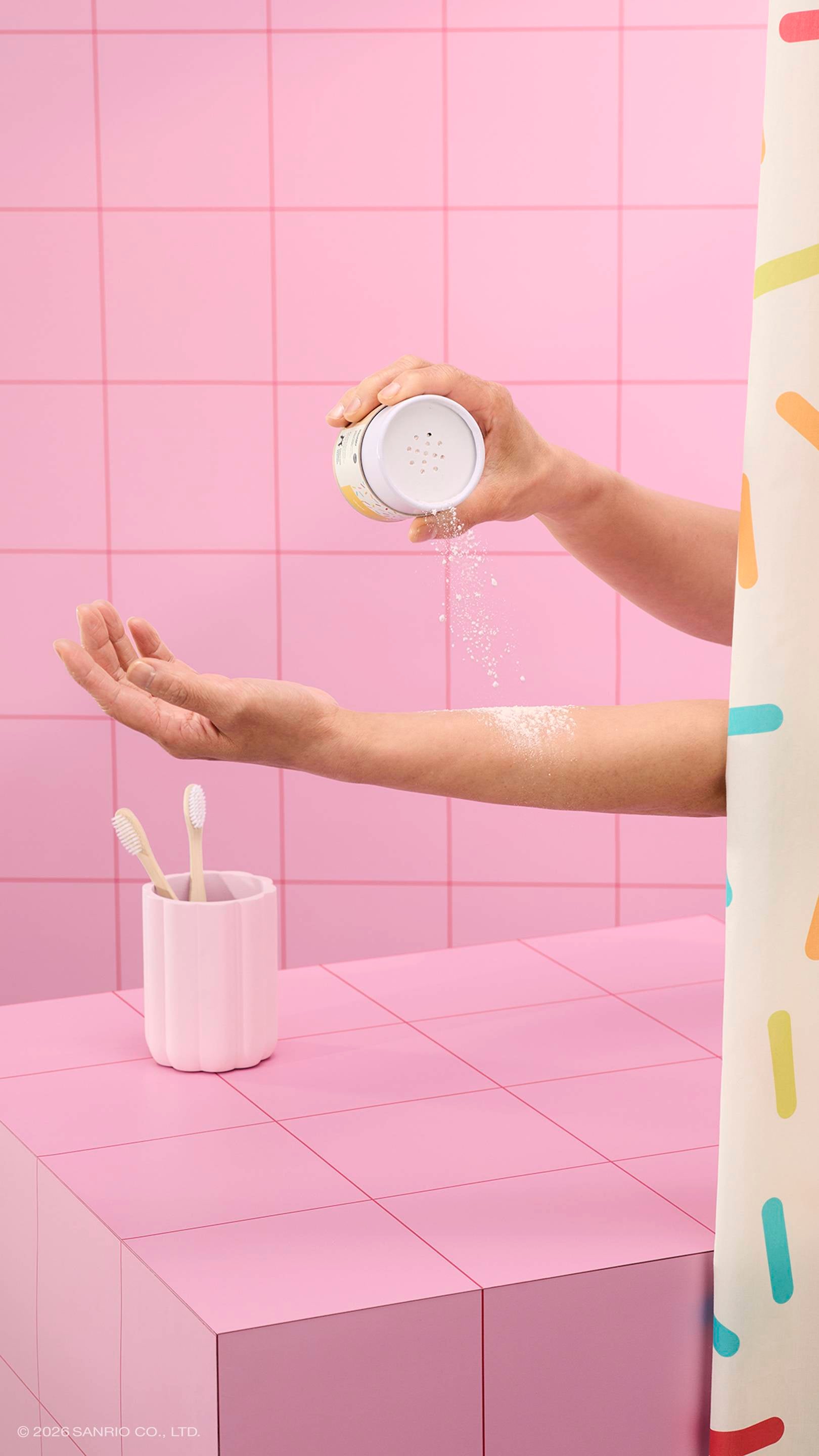 A hand shakes white dusting powder from a Lush x Pochacco tube onto another person's arm. The scene is set in a pink tiled bathroom with a sprinkle-patterned shower curtain and a toothbrush holder.
