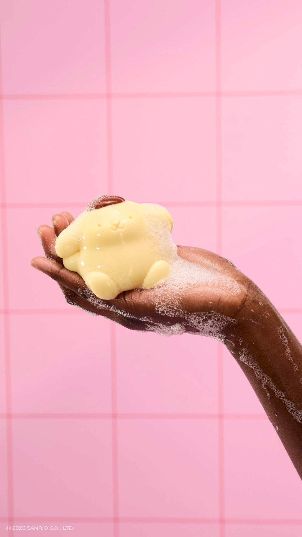A soapy hand holds a glossy, pale yellow Pompompurin shower jelly, covered in white bubbles. The scene is set against a background of pink square tiles.