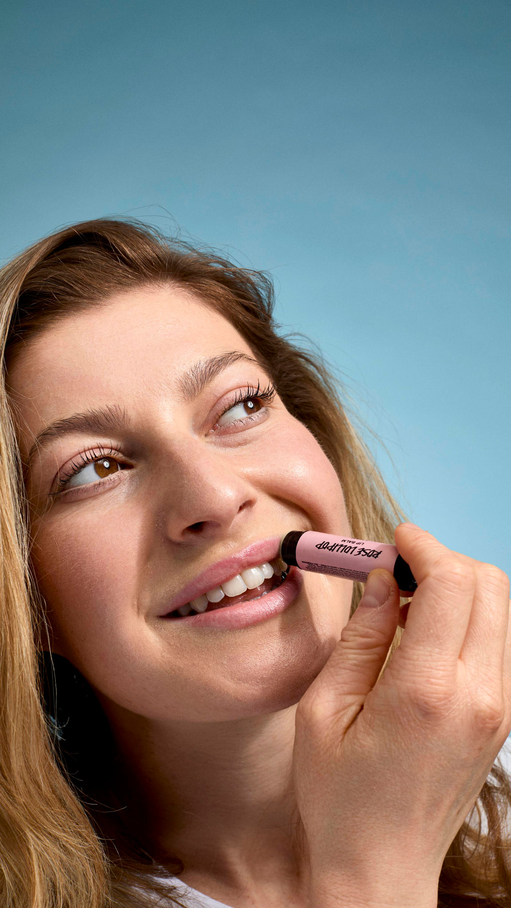 A close-up of a model applying 'Rose Lollipop' lip balm to their lips. They have a bright smile and are looking upwards and to the side, holding the pink-labelled Lush tube against their mouth against a light-blue background.