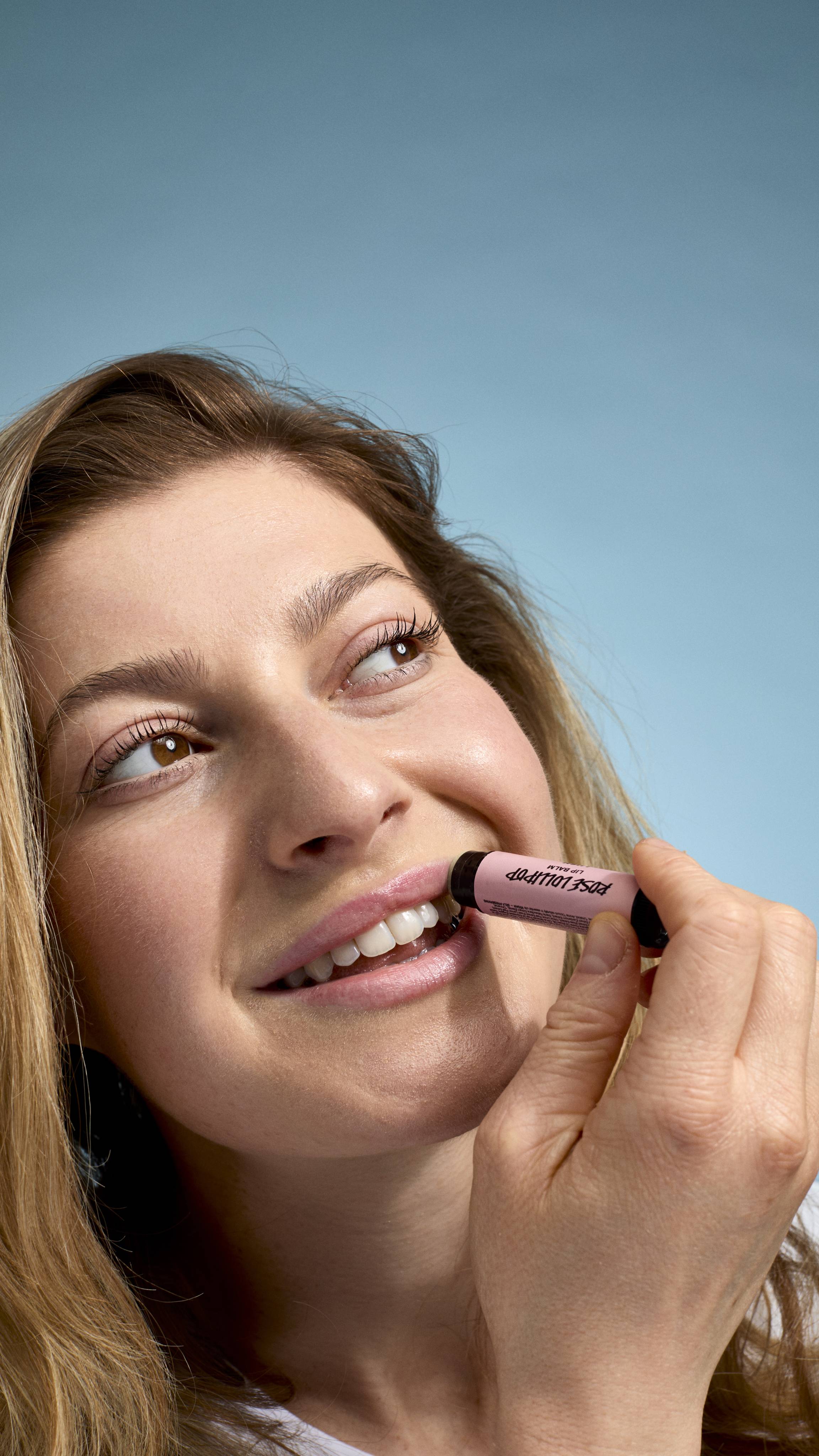 A close-up of a model applying 'Rose Lollipop' lip balm to their lips. They have a bright smile and are looking upwards and to the side, holding the pink-labelled Lush tube against their mouth against a light-blue background.