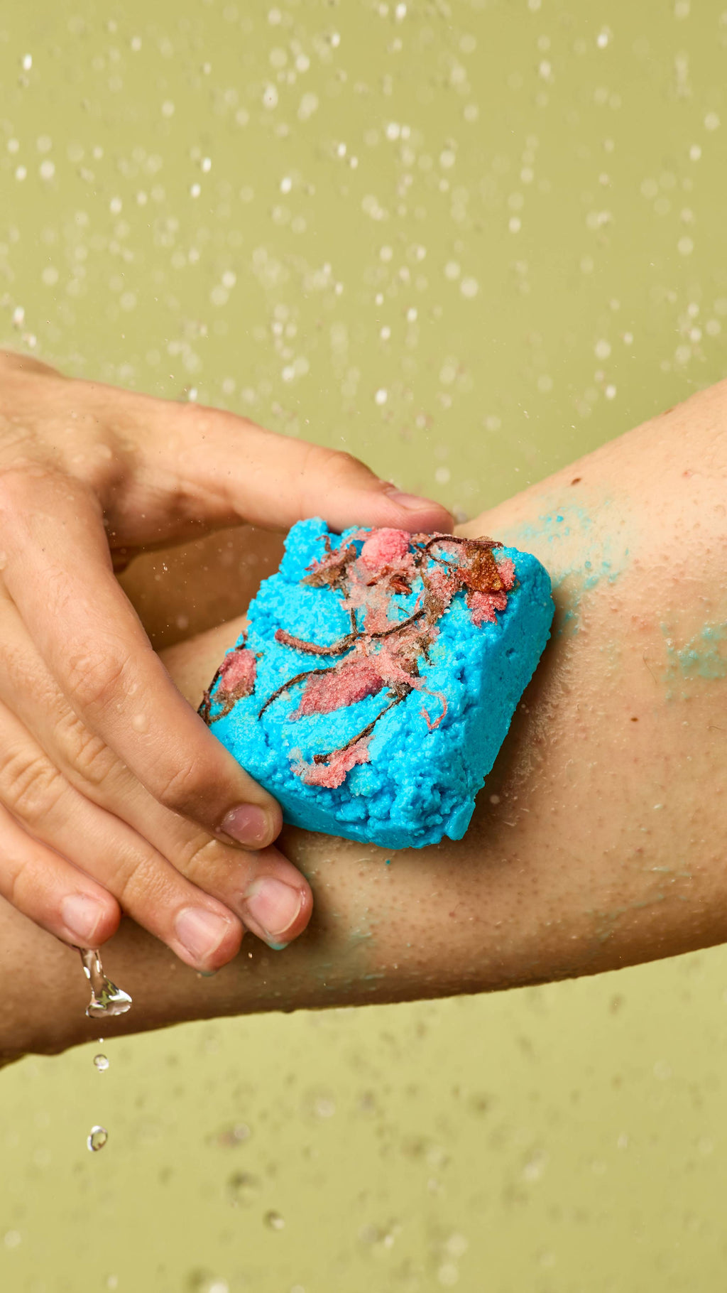 The image shows a hand rubbing the square, vibrant turquoise Rub Rub Rub solid body scrub, with a rough brown patch and salt crystals, onto an arm. Water droplets fall as blue-green residue forms on the skin, against a light green background.