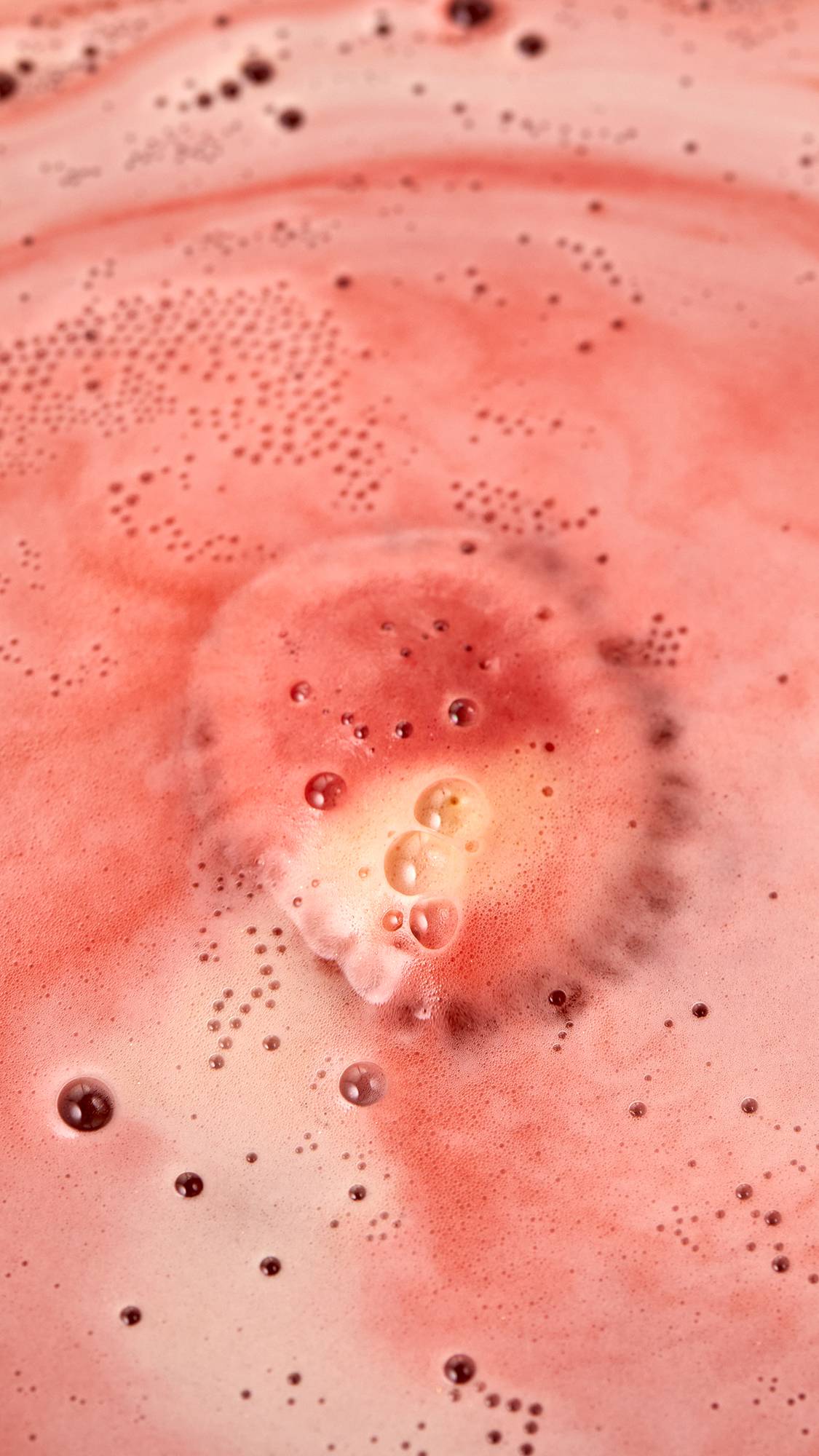 The image shows an aerial view of the Sticky Dates bath bomb as it starts to dissolve in the bath. The product is creating a thick blanket of warm, rich, rust-coloured, velvety foam with a hint of ivory foam in the centre.