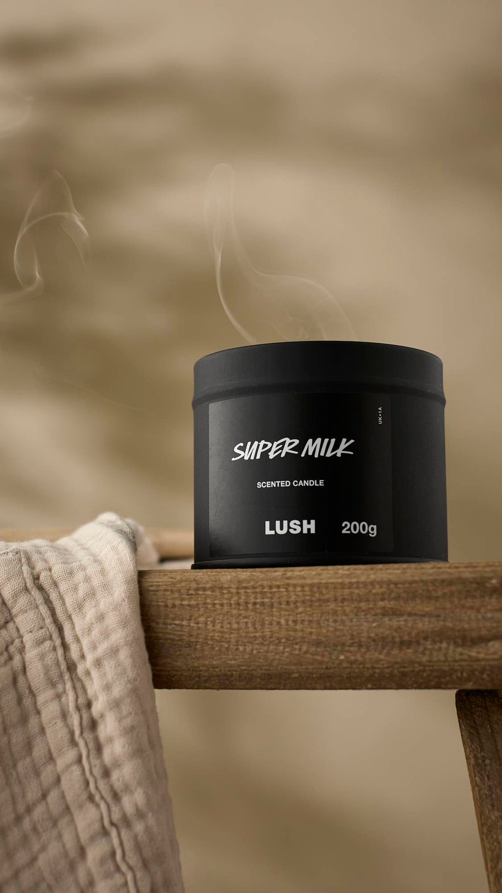 The matte black Super Milk Scented Candle is shown with its lid on, sitting on the edge of a rustic wooden frame or stool. A wisp of white smoke rises from the tin, and a light-coloured, textured cloth hangs in the foreground.