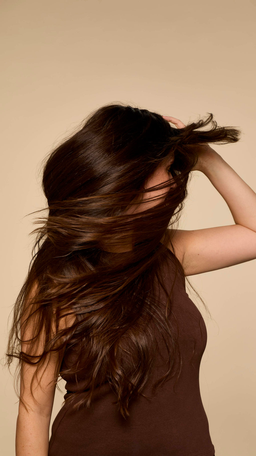 A person, seen in profile, with long, healthy dark hair is playfully tossing it to one side, creating a dynamic, windswept effect that showcases its volume and shine against a neutral background.