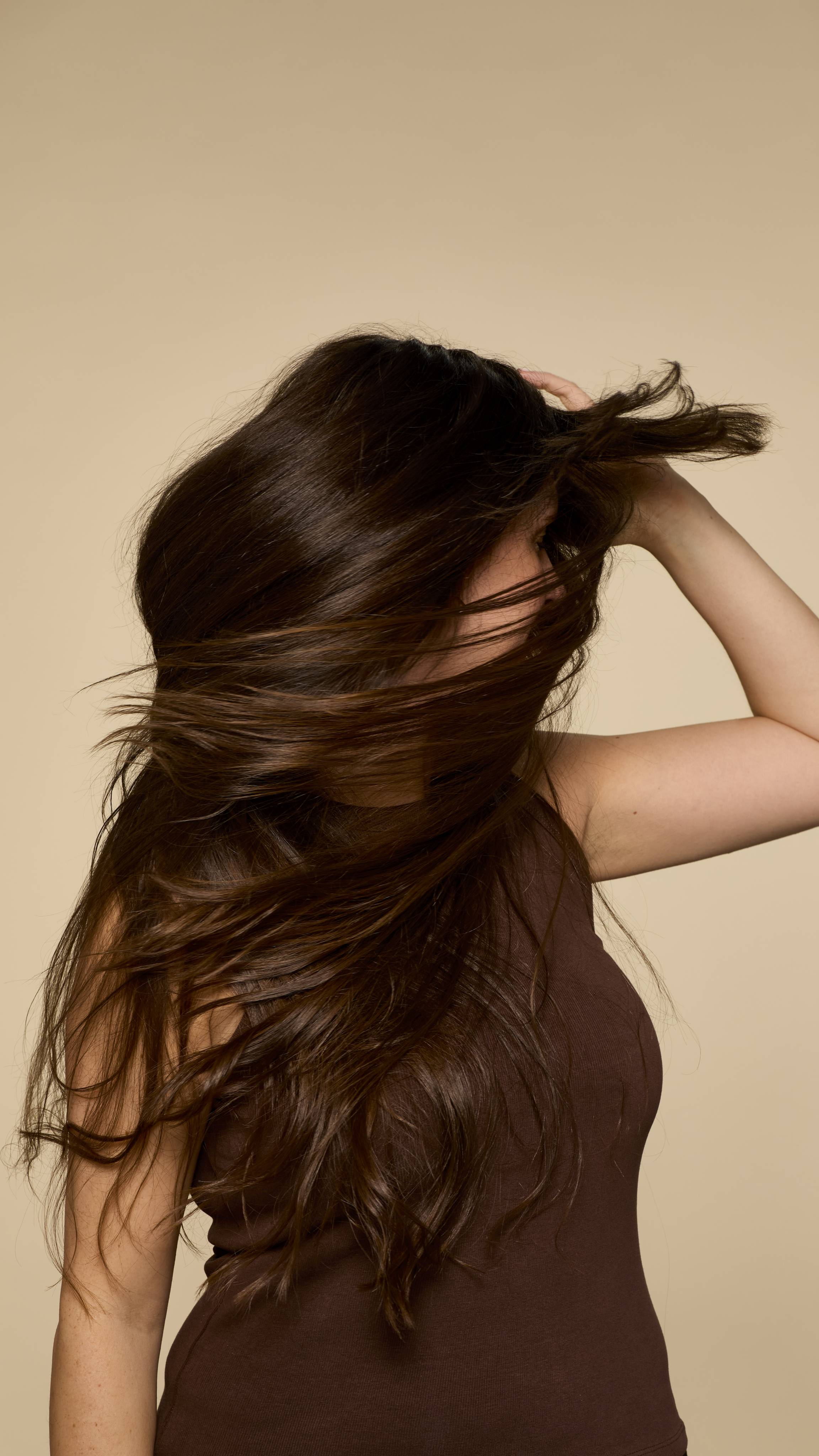 A person, seen in profile, with long, healthy dark hair is playfully tossing it to one side, creating a dynamic, windswept effect that showcases its volume and shine against a neutral background.