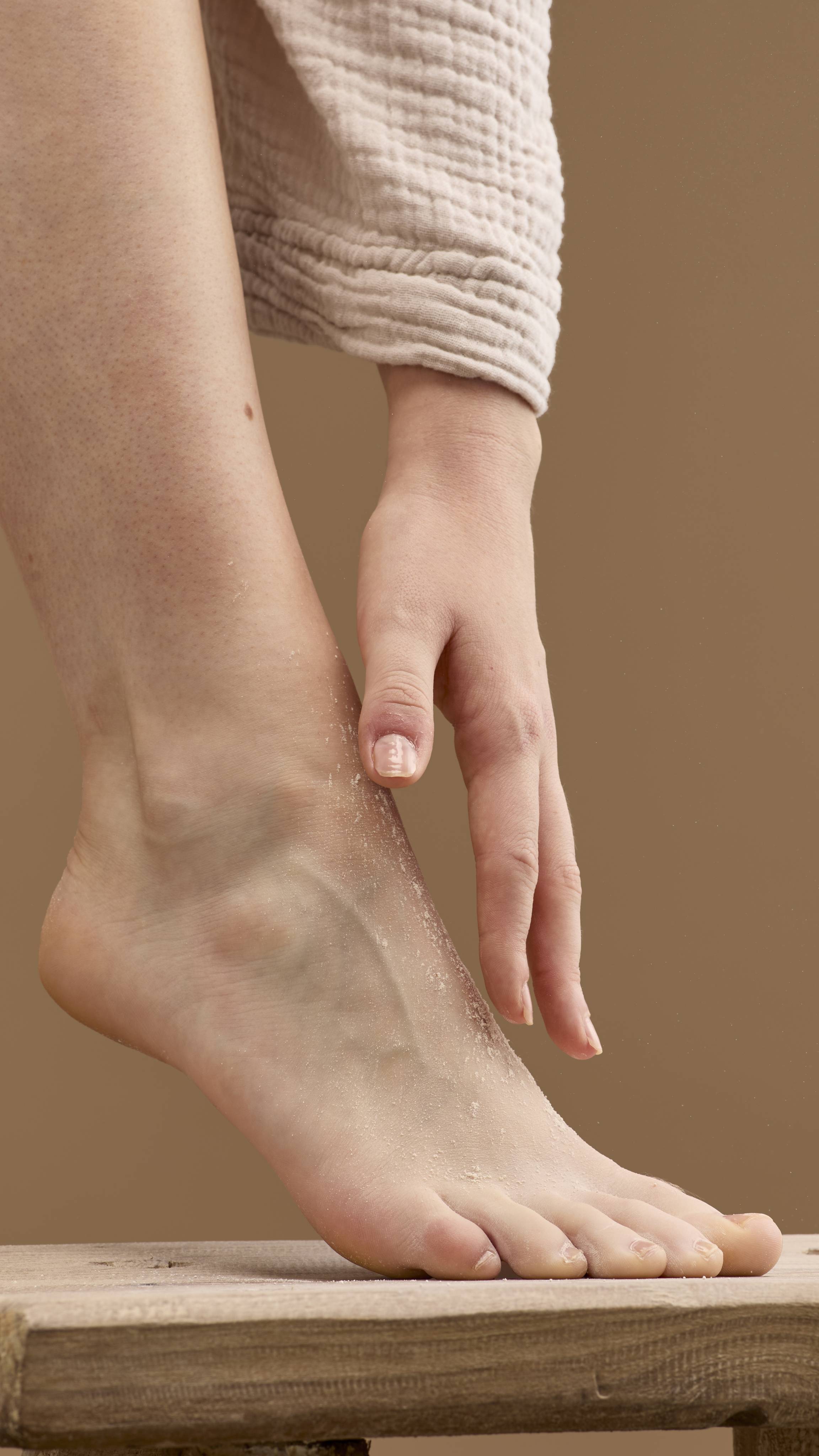 The image shows a hand rubbing a pale cream 'T For Toes' foot powder onto a bare foot and ankle. The foot rests on a wooden stool, with the person's robe visible against a solid beige background.