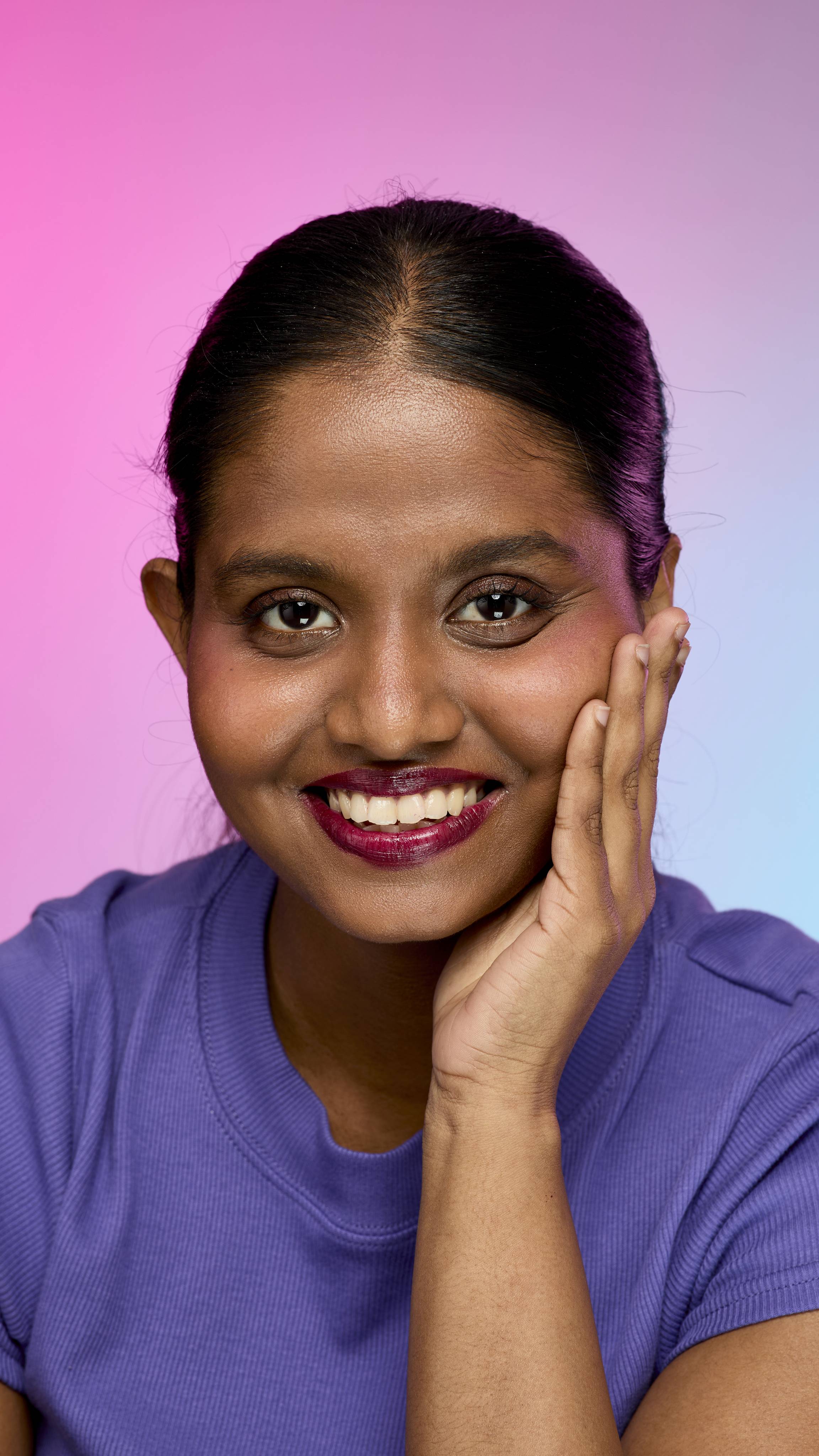 A smiling person with dark hair and skin, wearing a purple top, is shown from the chest up. They are wearing the Lush, deep plum Tartlet lip cream and have one hand touching the side of their face. The background is a pink and purple gradient.