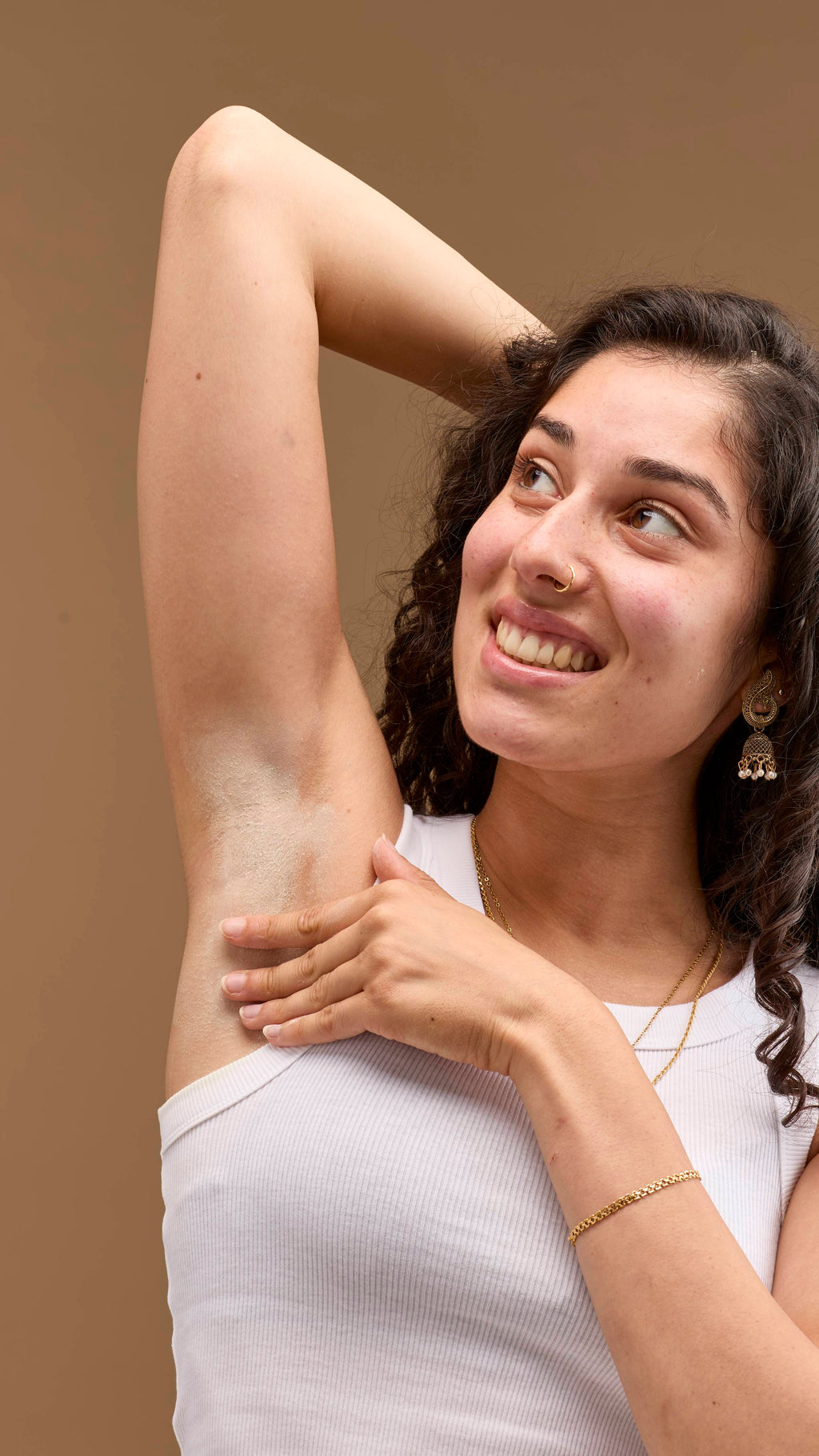 The image shows a person with dark, curly hair, smiling. They rub pale cream 'The Greeench' deodorant powder onto their raised underarm. The person wears a white vest top and necklaces against a light beige background.