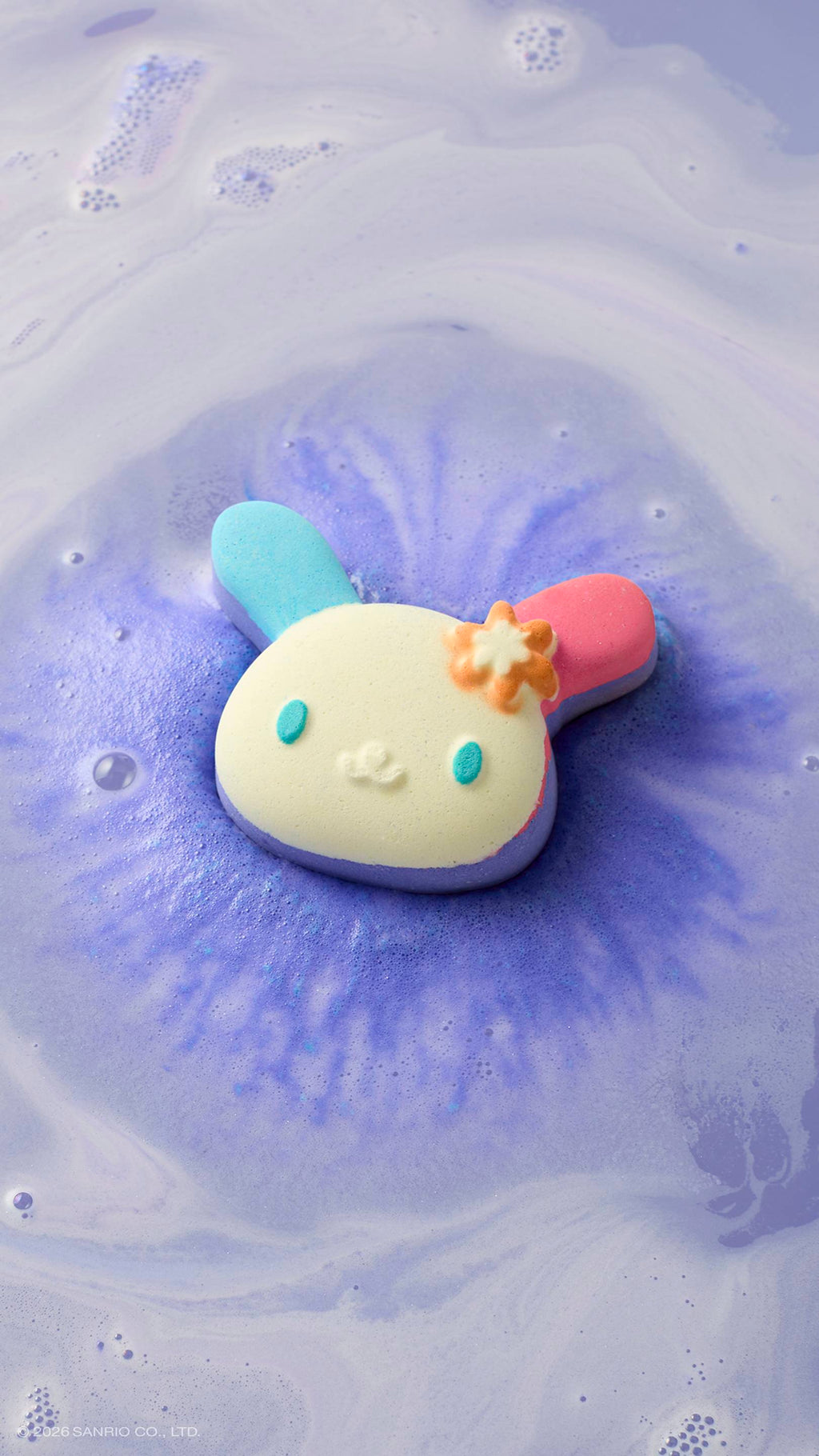 The bunny-shaped "U.SA.HA.NA" bath bomb with blue and pink ears floats in a bath of lavender-coloured water. It is surrounded by a vibrant purple fizz and swirls of white foam as it dissolves