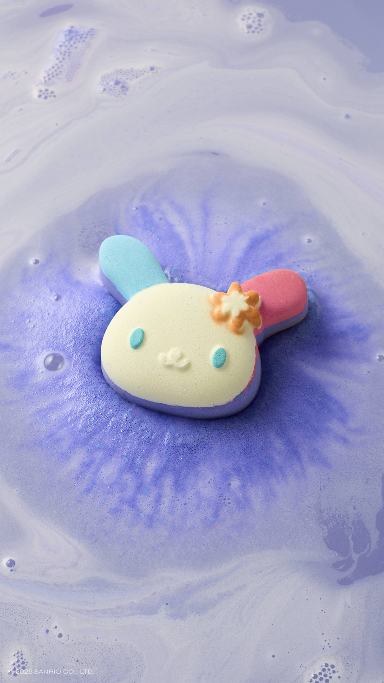 The bunny-shaped "U.SA.HA.NA" bath bomb with blue and pink ears floats in a bath of lavender-coloured water. It is surrounded by a vibrant purple fizz and swirls of white foam as it dissolves