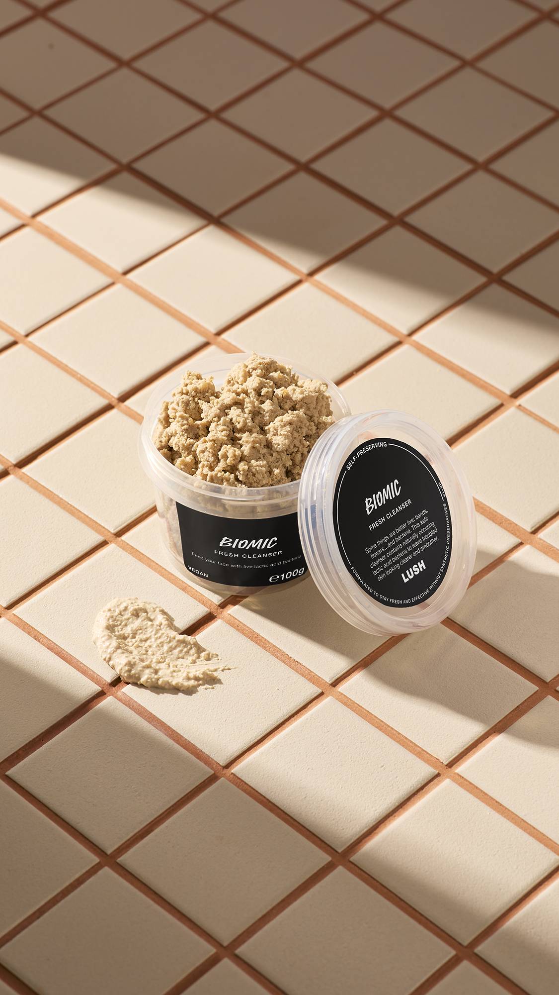 The image shows a clear, recycled plastic, open pot of Biomic facial cleanser with the lid leaning on the side and a smeared swatch of the textured product on the cream-tiled floor beside the pot.