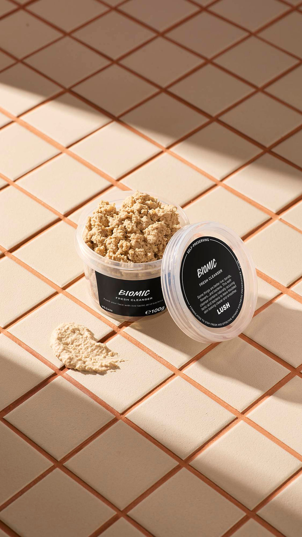 The image shows a clear, recycled plastic, open pot of Biomic facial cleanser with the lid leaning on the side and a smeared swatch of the textured product on the cream-tiled floor beside the pot.