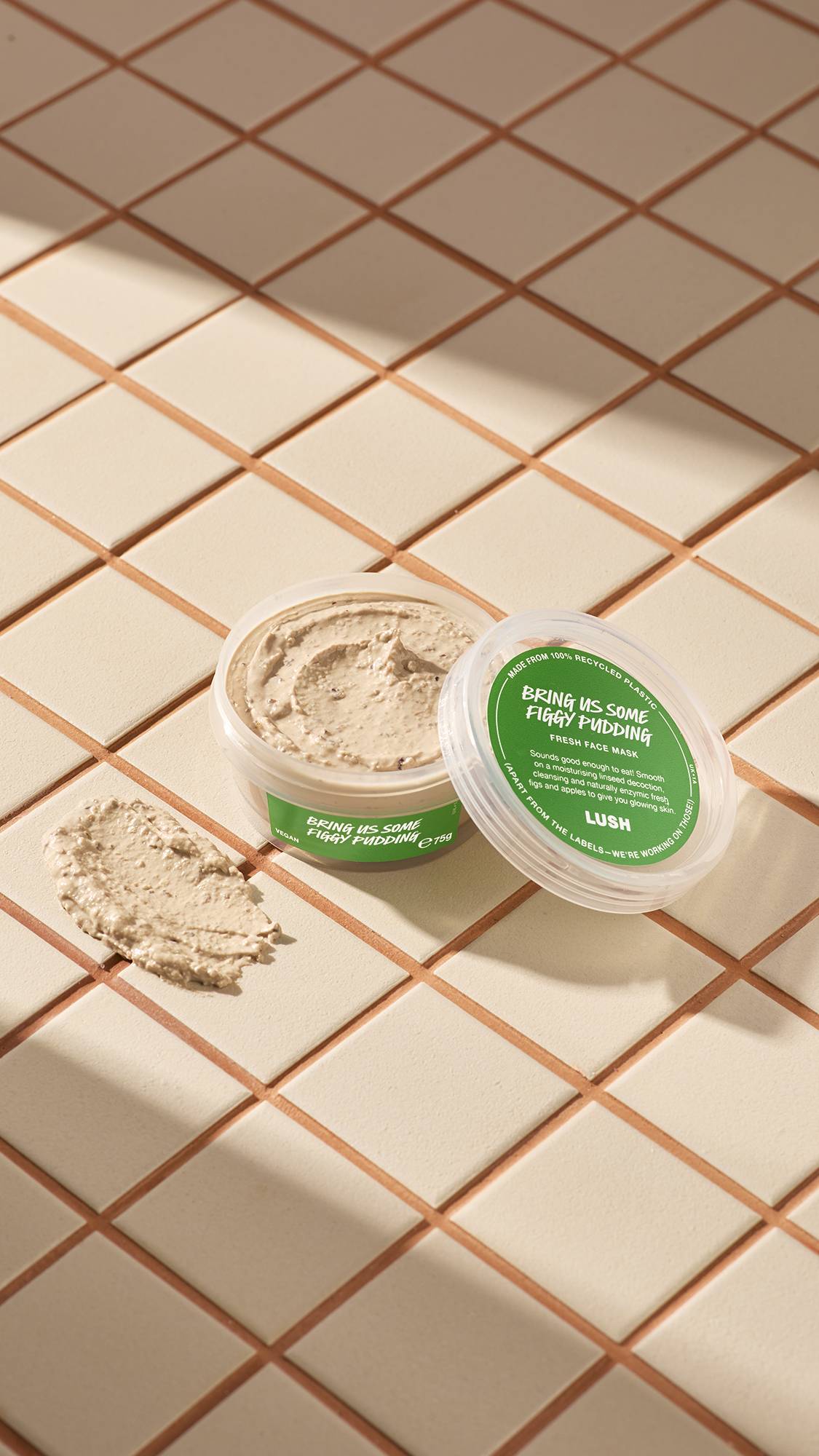 The image shows a clear, recycled plastic, open pot of Bring Us Some Figgy Pudding face mask with green labels, the lid leaning on the side and a smeared swatch of the textured product on the cream-tiled floor beside the pot.