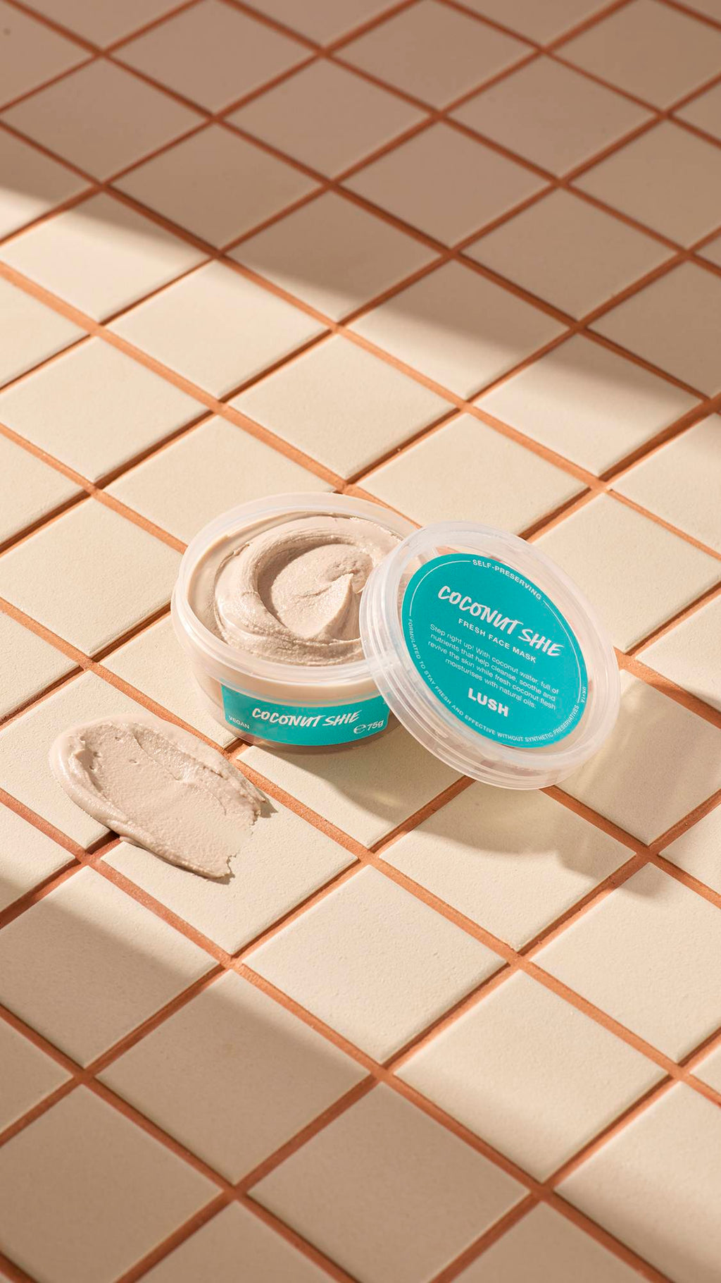 The image shows a clear, recycled plastic, open pot of Coconut Shie face mask with teal labels, the lid leaning on the side and a smeared swatch of the textured product on the cream-tiled floor beside the pot.