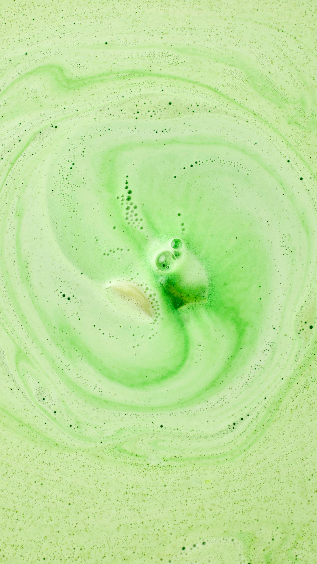 A top-down view captures the Cold Bath Bomb dissolving in the water, creating a mesmerizing swirl of frothy green and creamy white foam.